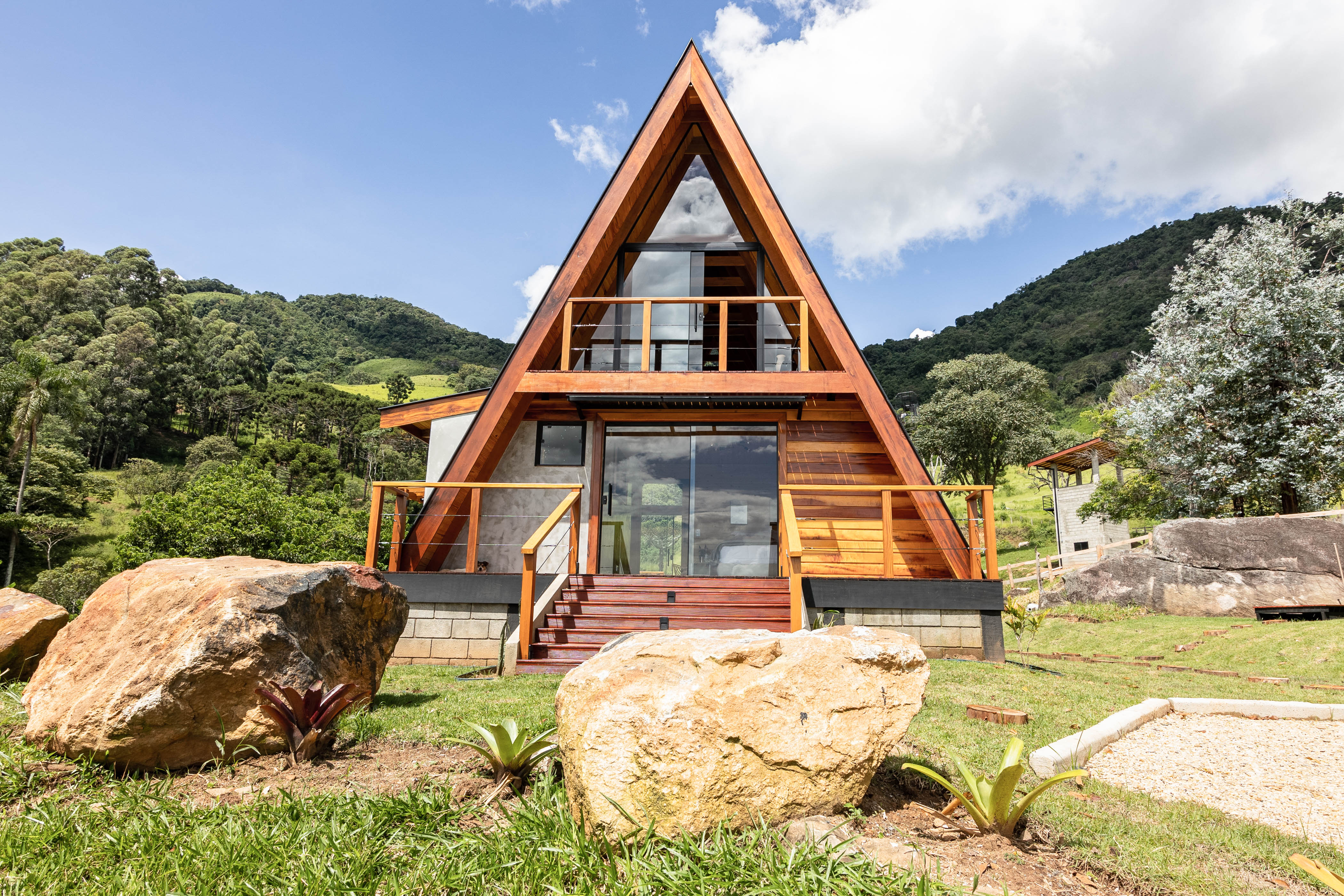 A modern A-frame house made of wood and glass, set against a backdrop of lush green hills and blue sky. The house features a large front balcony, a wooden staircase leading to the entrance, and large windows reflecting the surrounding nature. In the foreground, there are large rocks and small plants, adding to the natural landscape.