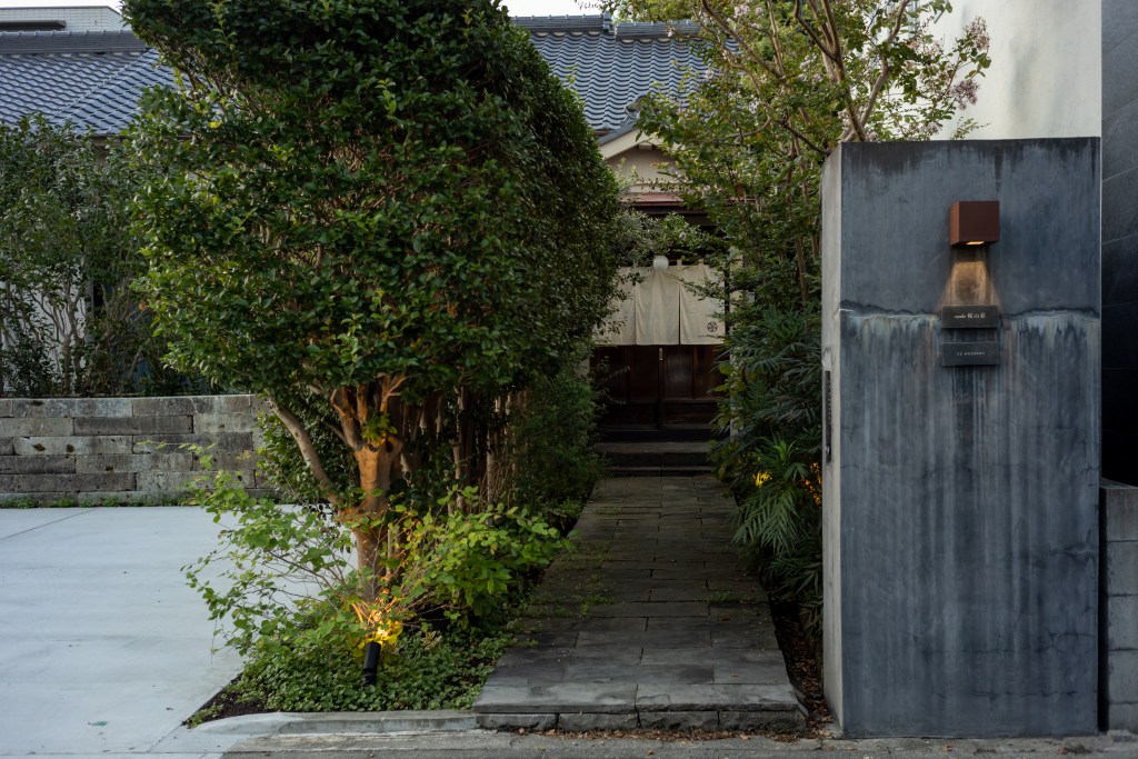 A serene entrance pathway lined with lush greenery leads to a traditional style wooden door, partially obscured by a fabric curtain. The entrance is framed by well-trimmed hedges and illuminated by soft outdoor lighting, inviting guests into a peaceful setting.