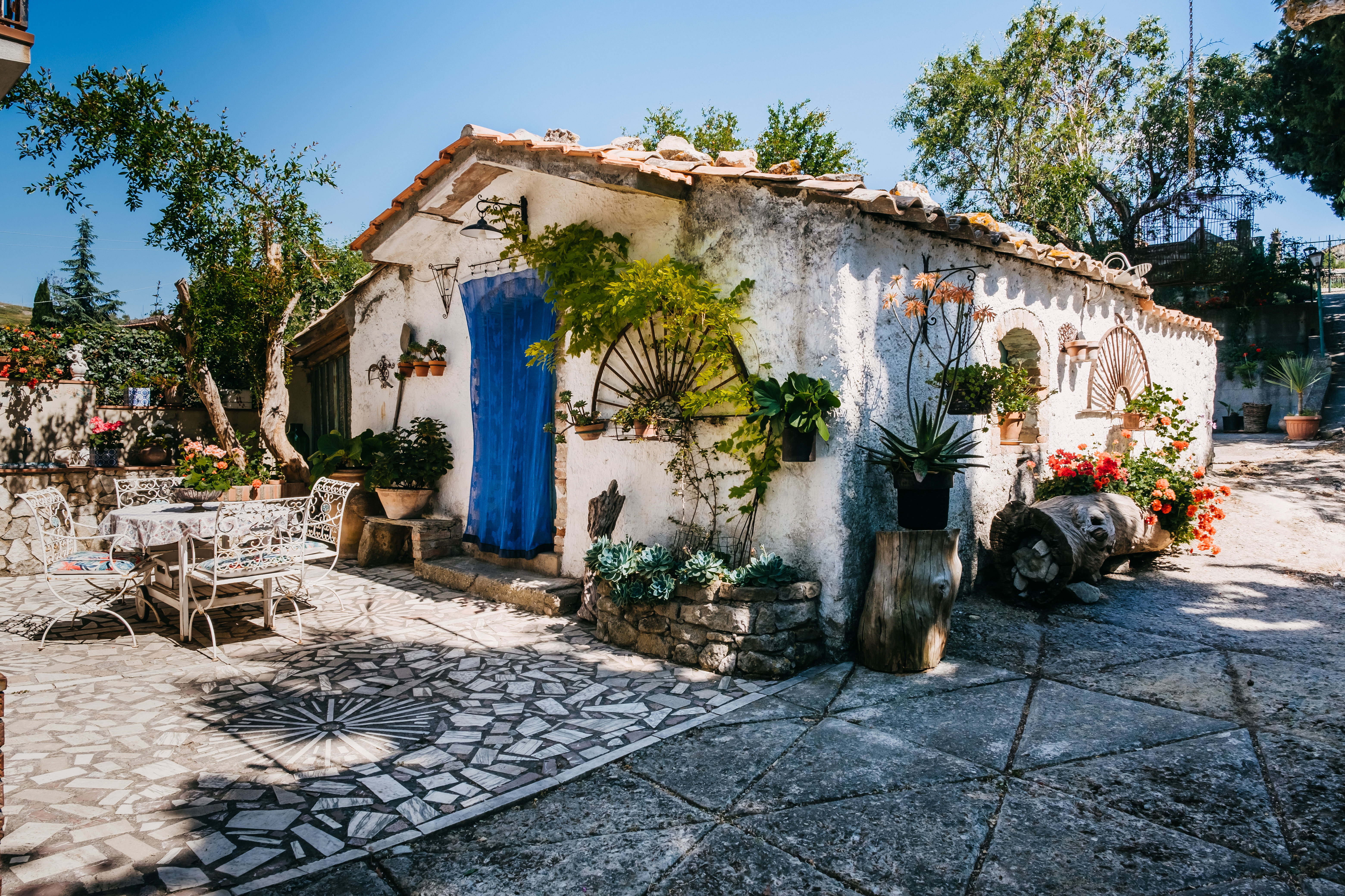 An Italian style villa surrounded by lush greenery and flowers. It has a blue tapestry on its door and faces a stone patio.