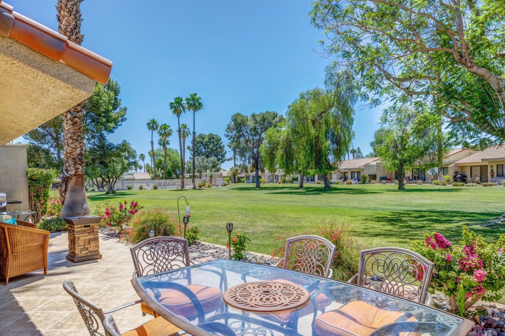  A sunny outdoor patio area featuring a glass dining table with metal chairs and cushions. In the background, there is a lush green lawn with various trees, including palm trees, and several houses visible in the distance. The sky is clear and blue.