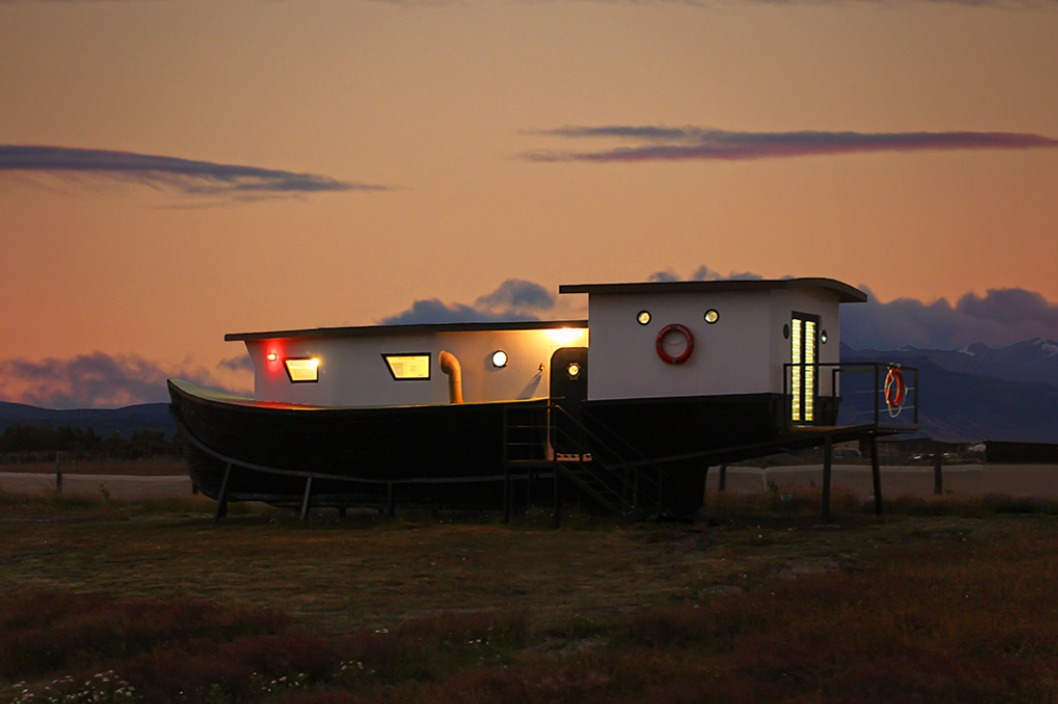 A unique, modern house designed to resemble a boat, illuminated at dusk. The structure features large windows, a life preserver on the side, and is set against a backdrop of mountains and a colorful evening sky.