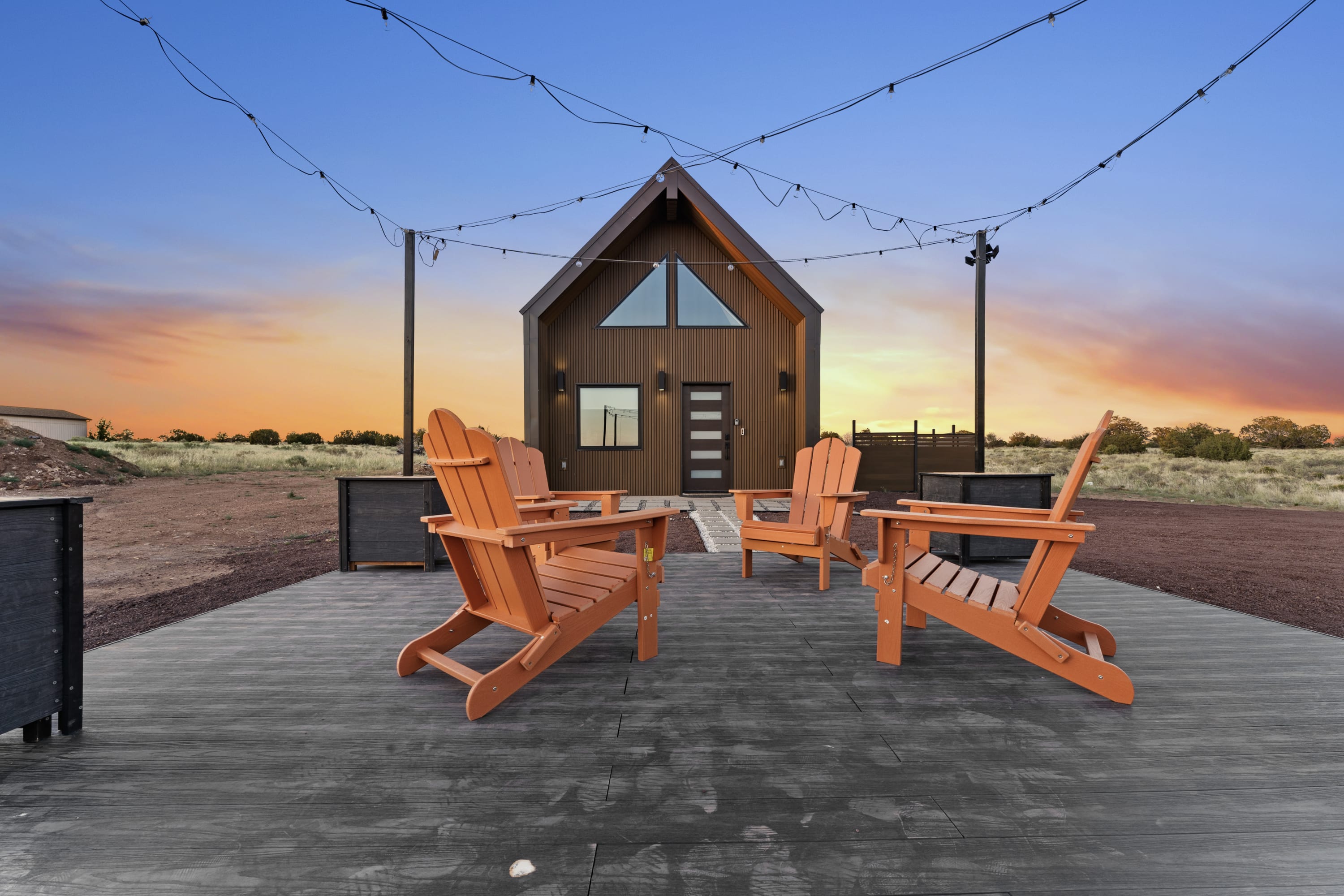 Outdoor seating area with four wooden chairs in front of a modern, brown A-frame cabin at sunset.