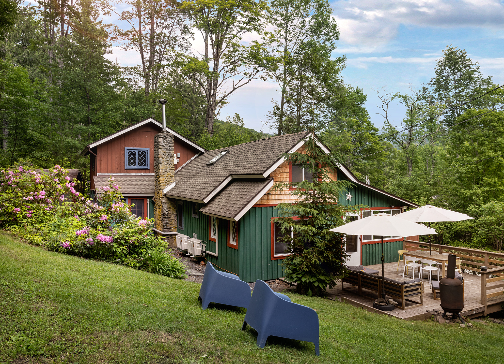 Charming cabin with a green and brown facade, surrounded by lush trees and vibrant flowers, featuring a patio with outdoor seating and an umbrella.