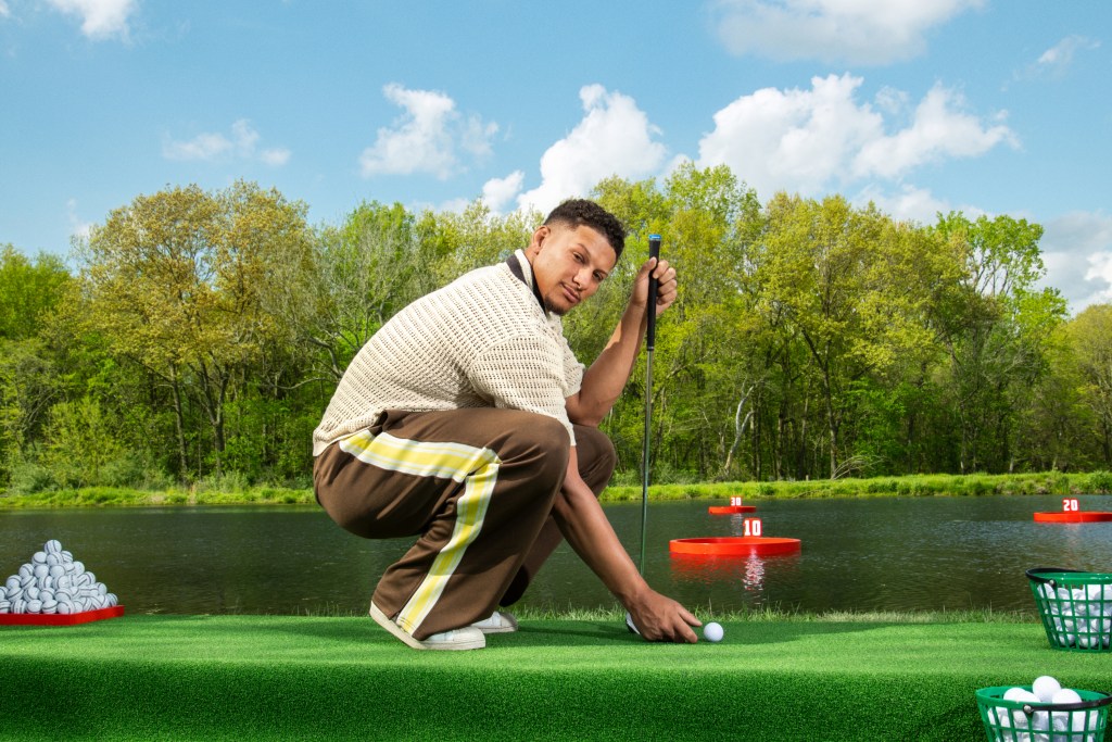 Person crouching on a putting green by a lake, preparing to hit a golf ball towards floating red targets.