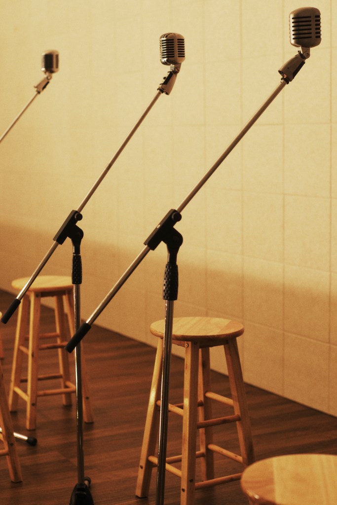Three vintage microphones on stands in front of wooden stools in a dimly lit room.