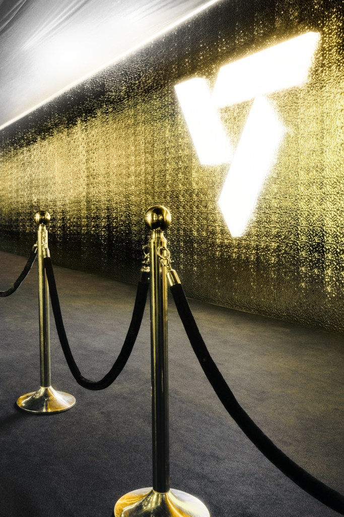 Artistic glowing symbol on a shimmering gold wall with velvet rope barriers.
