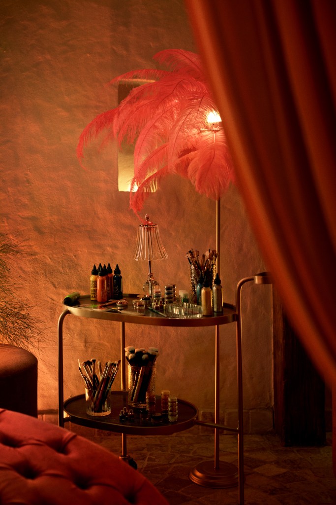 A cozy and luxurious corner with a pink feather lamp, makeup supplies, and art materials arranged neatly on a gold trolley, set against a textured wall in soft lighting.