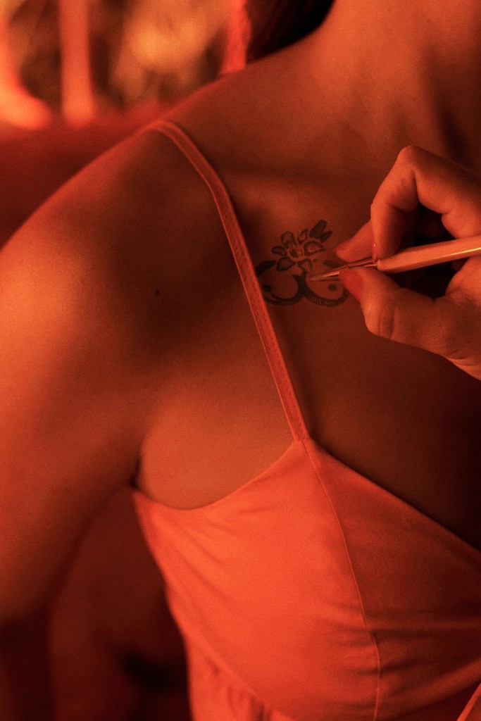 A close-up of a person wearing an orange dress, with a hand drawing a temporary tattoo of an Om symbol and floral design on their shoulder under warm, ambient lighting.