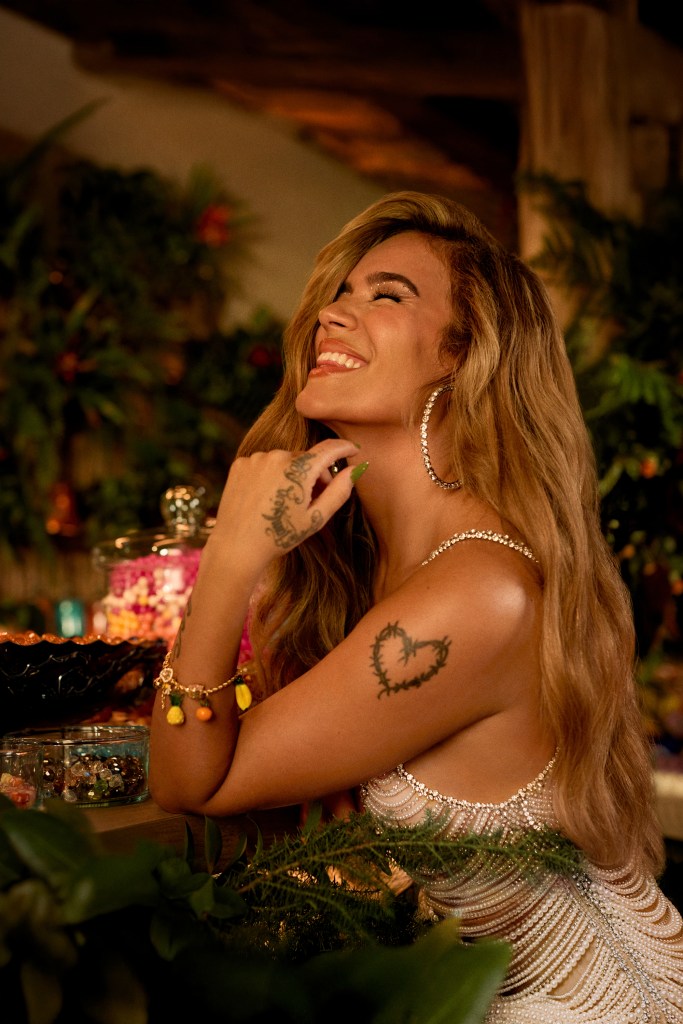 A close-up of a person with long blonde hair and elegant jewelry, wearing a beaded dress, sitting in a cozy, warmly-lit space surrounded by greenery and colorful candy jars.