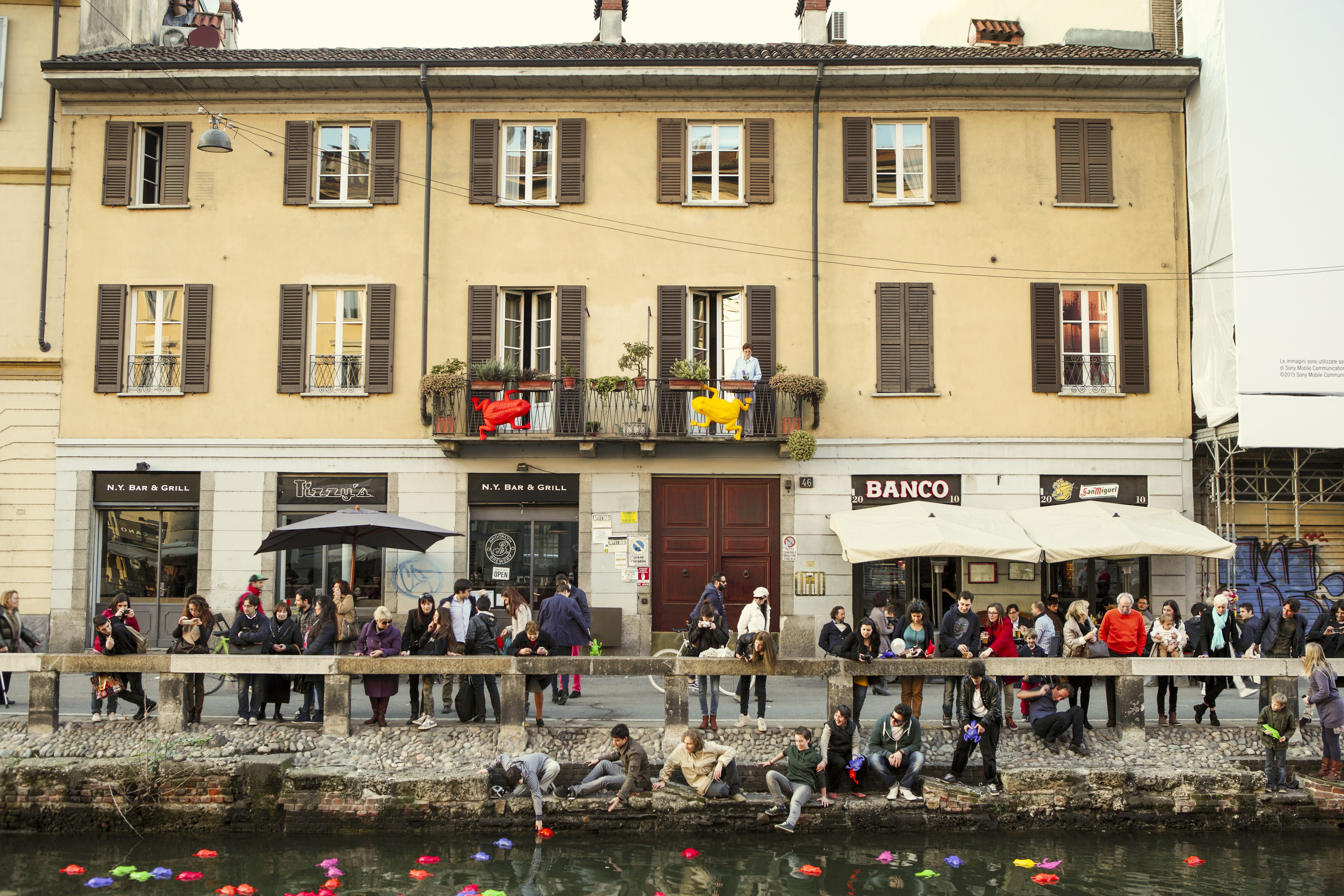 Group of people gathered by a canal with colorful floating decorations, in front of a yellow building with restaurants and balconies adorned with plants.