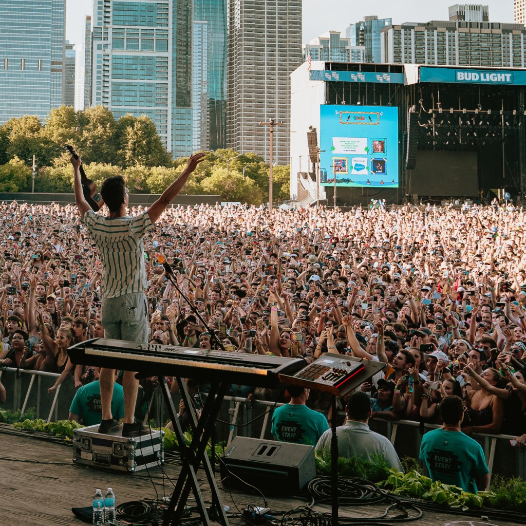 A performer with arms raised stands on a stage overlooking a cheering festival audience, with a blue event screen and city buildings visible in the distance.