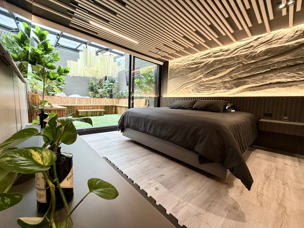 Modern bedroom with a large glass window overlooking a lush garden, featuring a textured wall, ceiling slats, and a black bedspread.