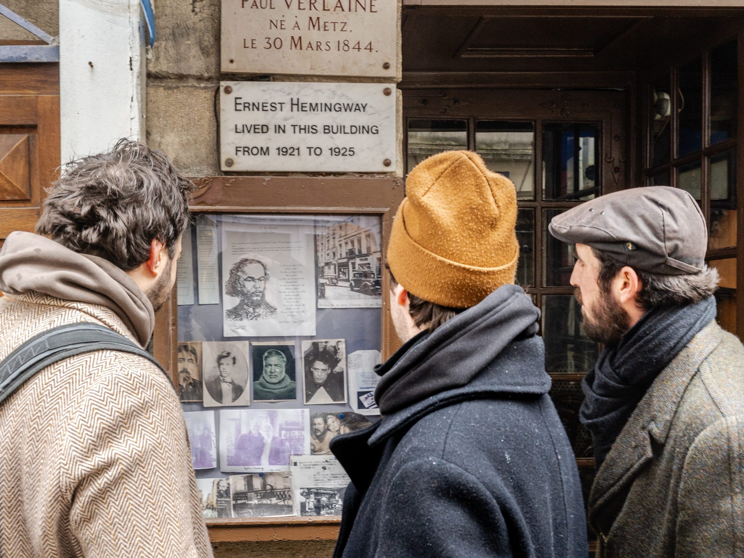 Three people in winter coats and hats stand closely together, looking at historical plaques and vintage photographs displayed on a building wall.