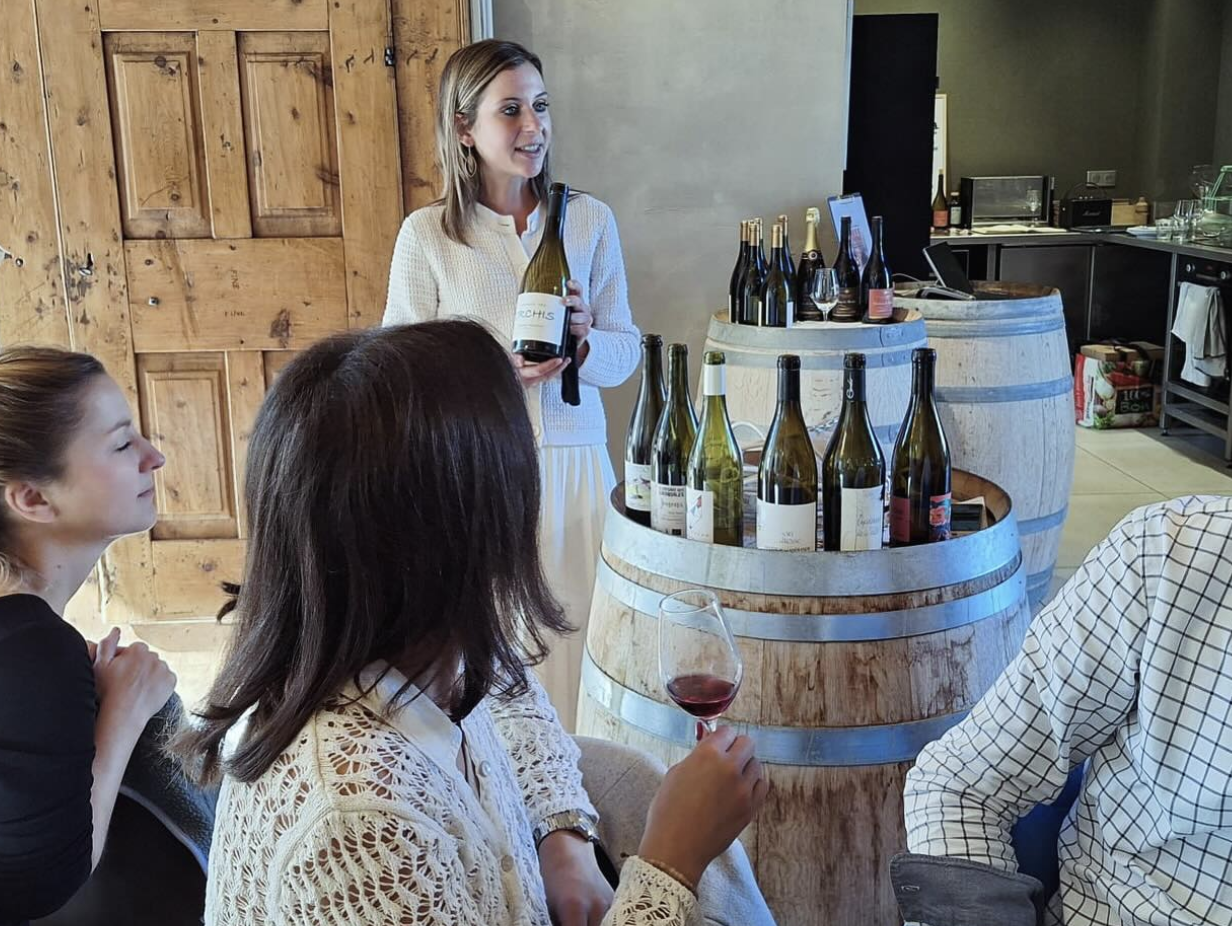 A small group participates in a wine tasting session, with bottles displayed on barrels and one person presenting a wine.