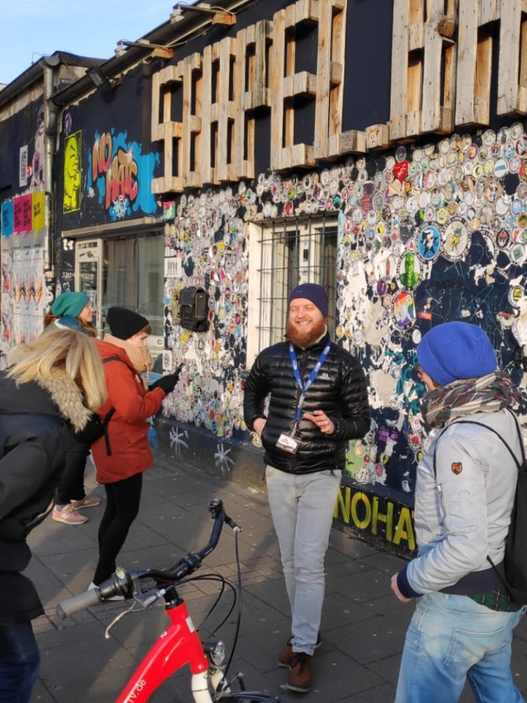 Group of people gathered outside a graffiti-covered building with a wooden 'Street Art' sign, participating in a street art tour on a sunny day.