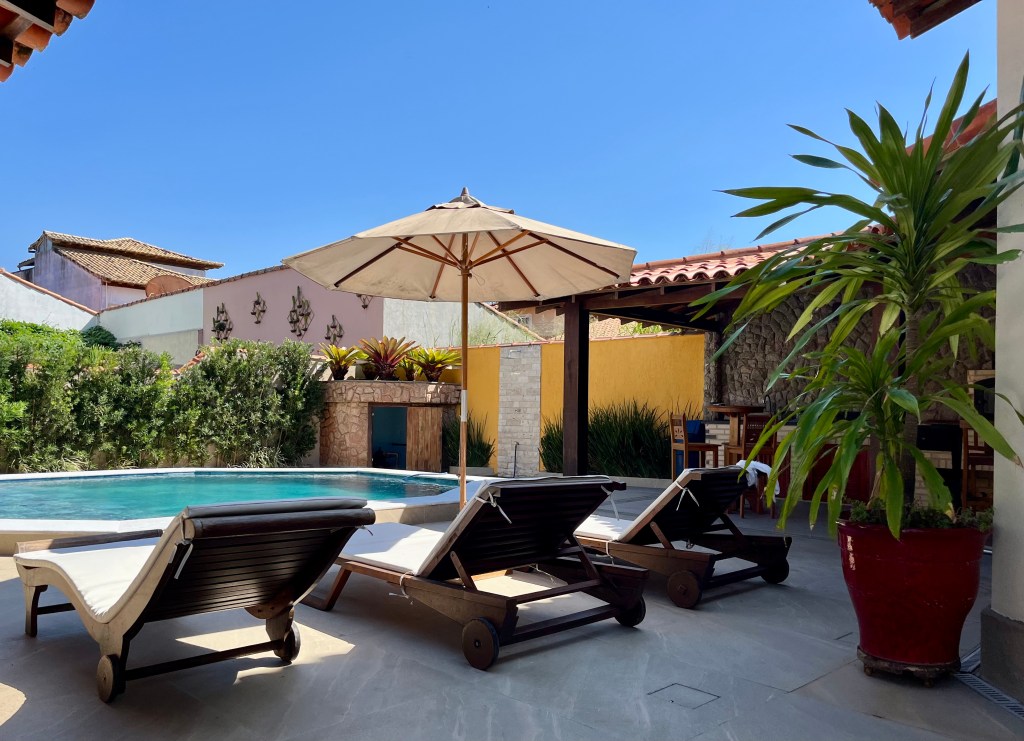 Outdoor patio with a swimming pool, surrounded by lounge chairs and a large umbrella under clear blue skies, with vibrant plants and colorful walls in the background.