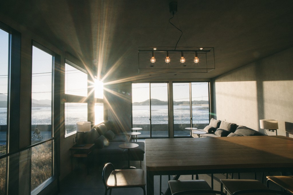 A modern, sunlit living space with large windows overlooking a serene waterfront and distant mountains during sunset.