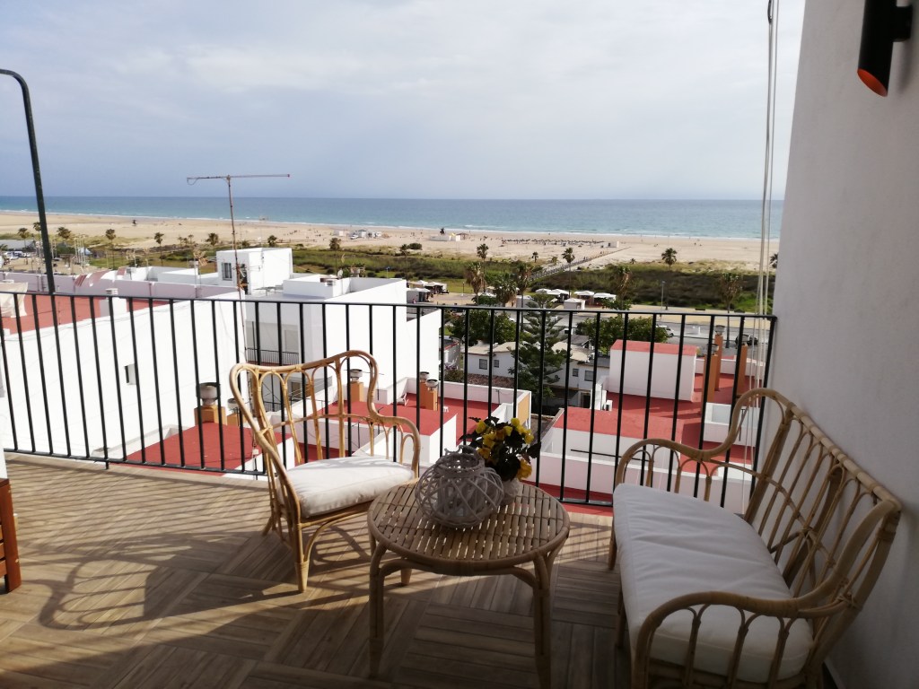 Coastal balcony with wooden wicker furniture overlooking rooftops, a sandy beach, and the blue ocean under a partly cloudy sky.