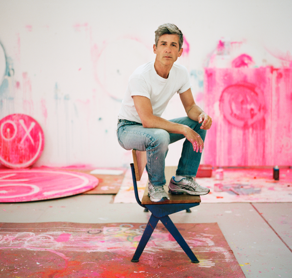 A casually dressed individual sits on a modern stool surrounded by a studio filled with pink art and paint splatters.