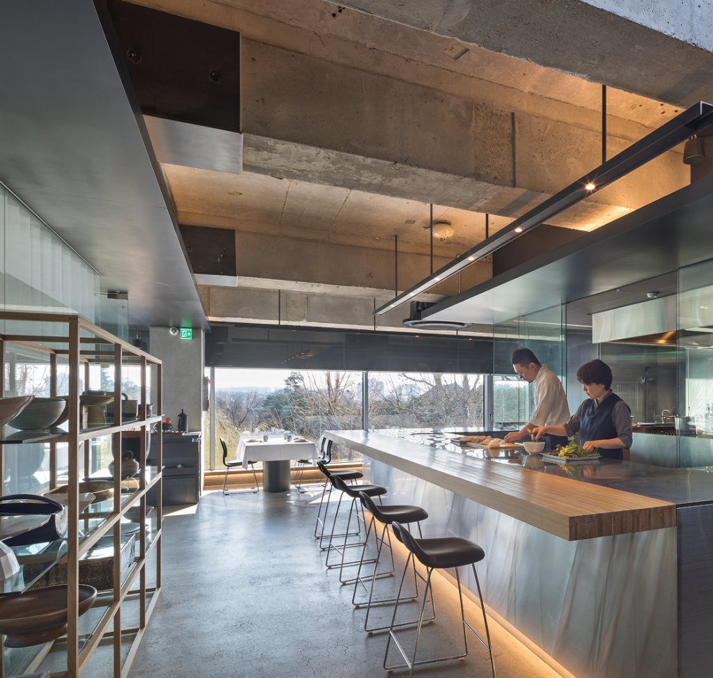 Two chefs prepare food together in a sleek, contemporary kitchen with large windows overlooking nature.