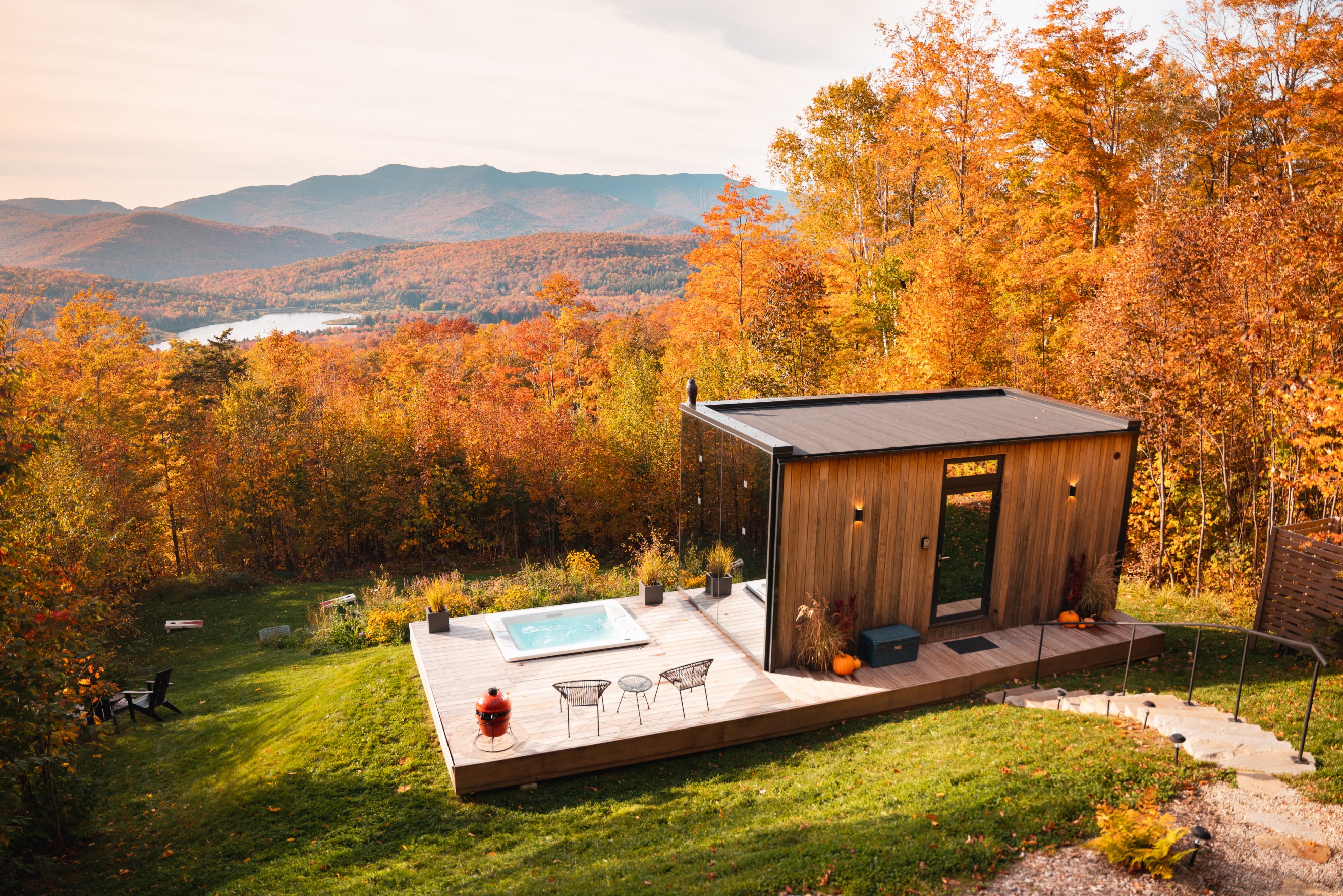 A modern wooden cabin with a deck and hot tub sits amidst vibrant autumn foliage overlooking a scenic mountain and lake view.