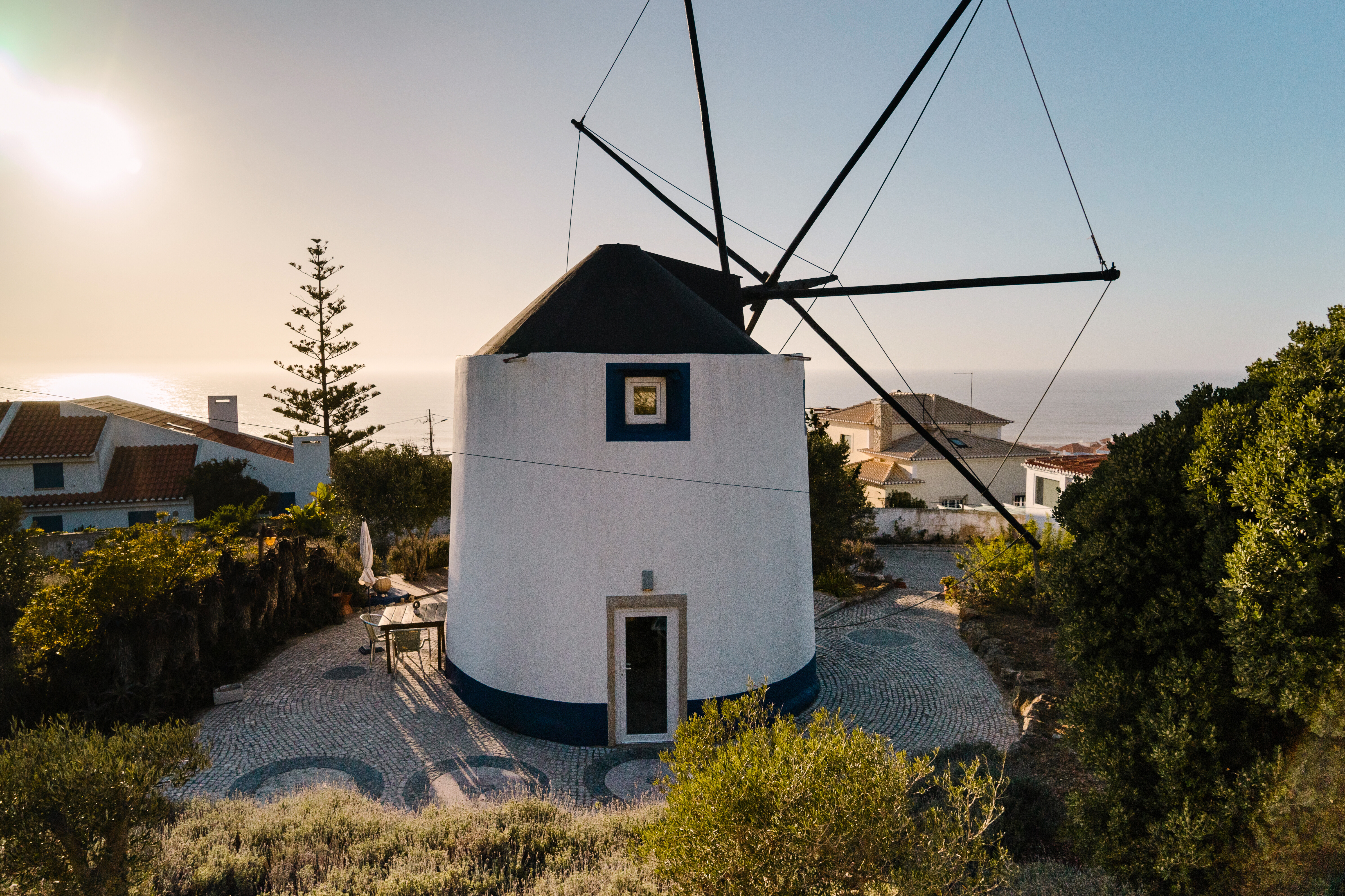A charming, round white windmill stands on a stone courtyard overlooking the ocean at sunset.