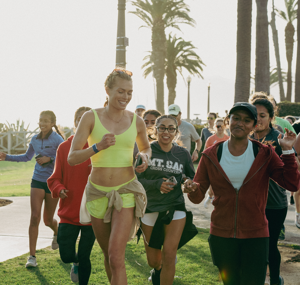 A group of people jog together outdoors along a palm-lined path on a sunny day.
