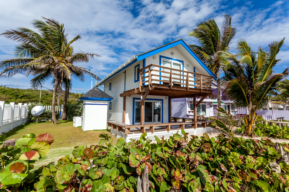 A coastal house with blue trim and a spacious balcony is surrounded by palm trees and tropical plants under a sunny sky.