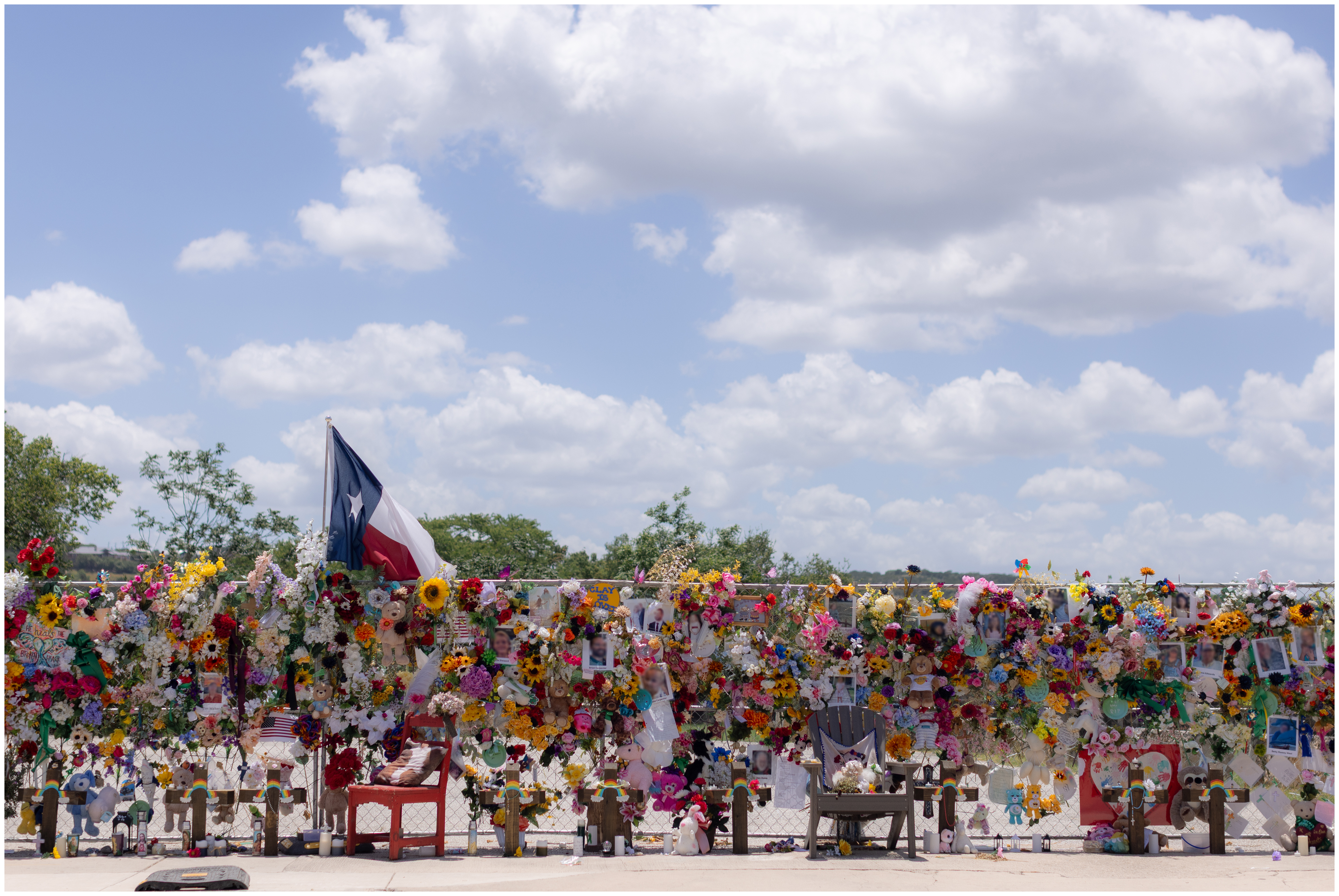 A memorial of flowers and stuffed animals tucked into a chain link fence, with a blue sky and white clouds in the background. A Texas flag flies above the fence.