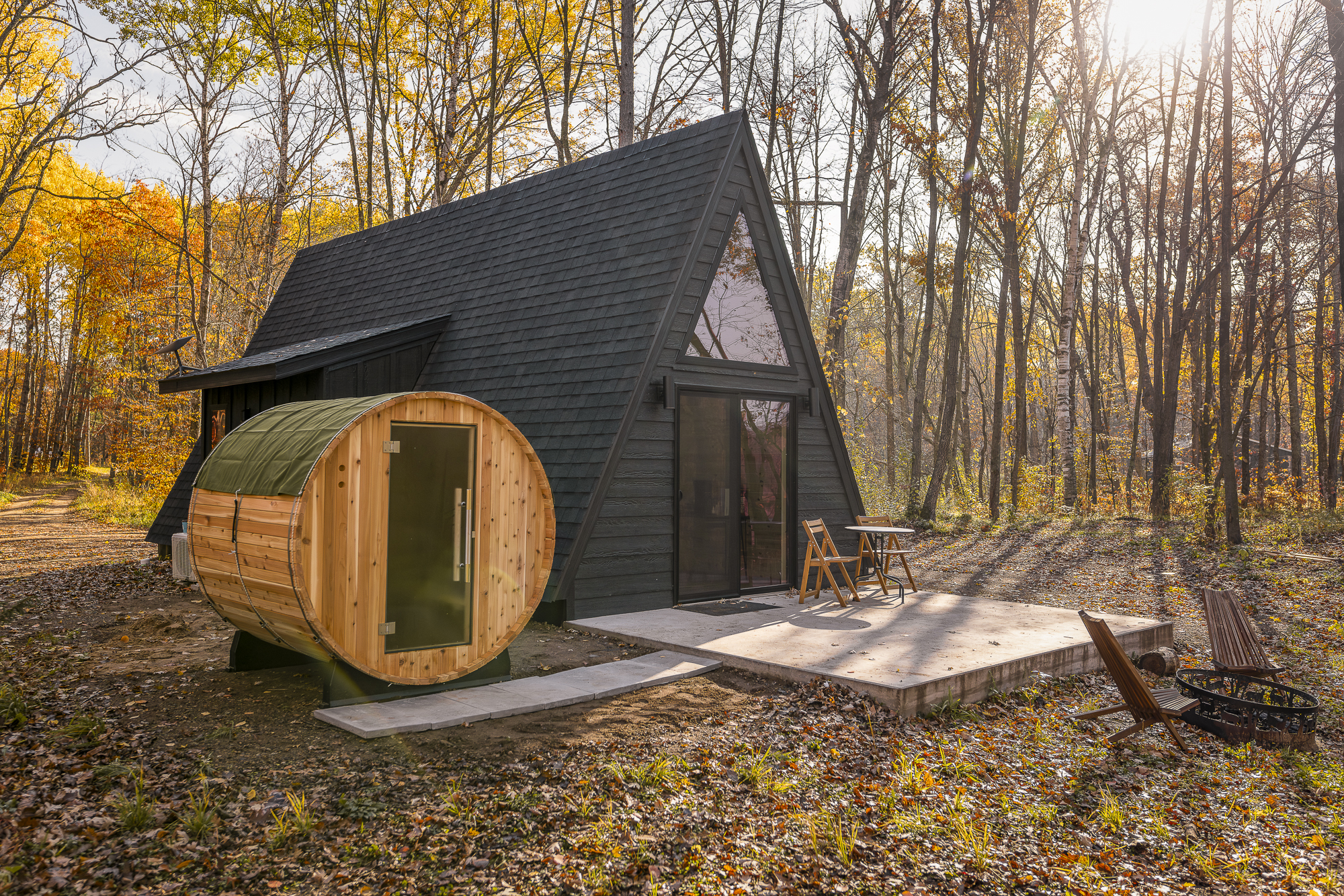 A modern black A-frame cabin with a barrel sauna sits in a sun-dappled clearing surrounded by trees.