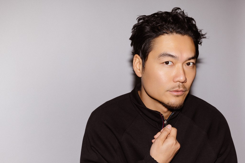 Person wearing a black zip-up sweater against a neutral gray background, holding the collar of the sweater with one hand.