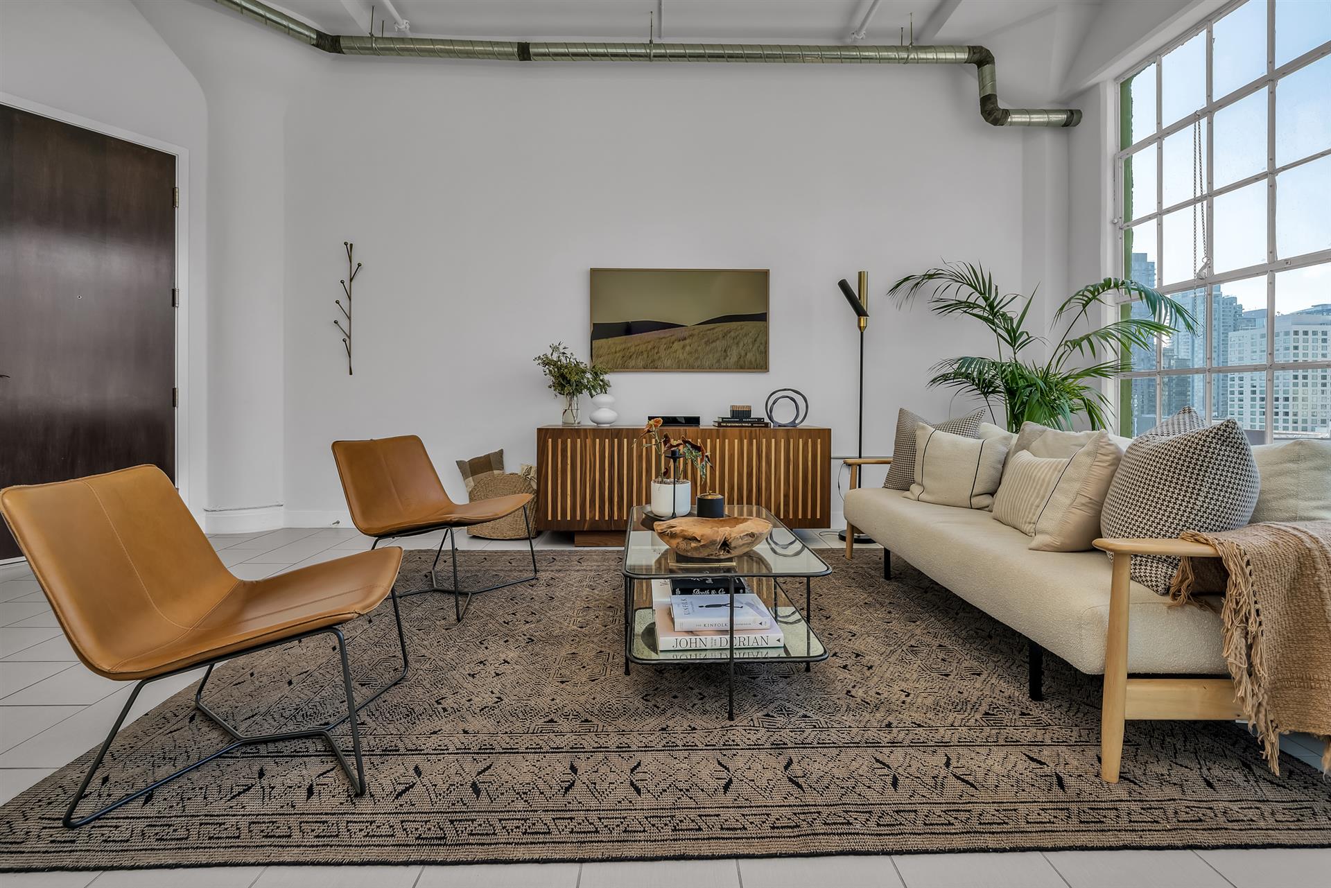 Bright and modern living room with leather chairs, a beige sofa, a patterned rug, and a large window offering city views.