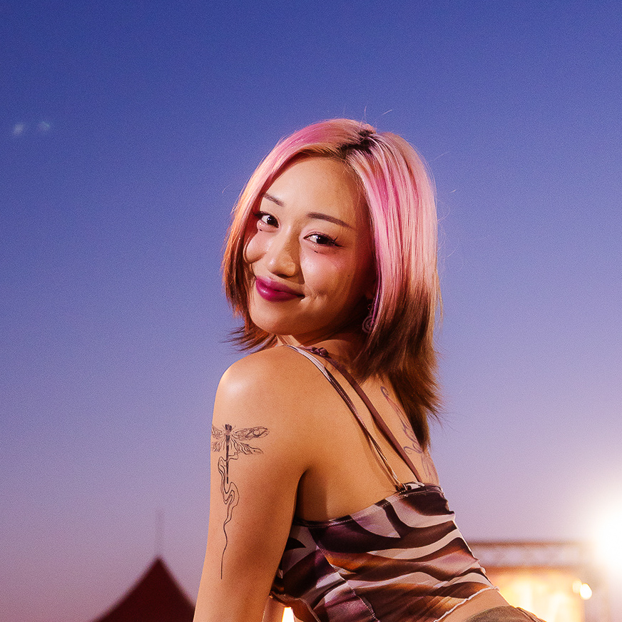 A person with pink and brown hair wearing a patterned top stands against a twilight sky, with a visible tattoo of a dragonfly on their shoulder.