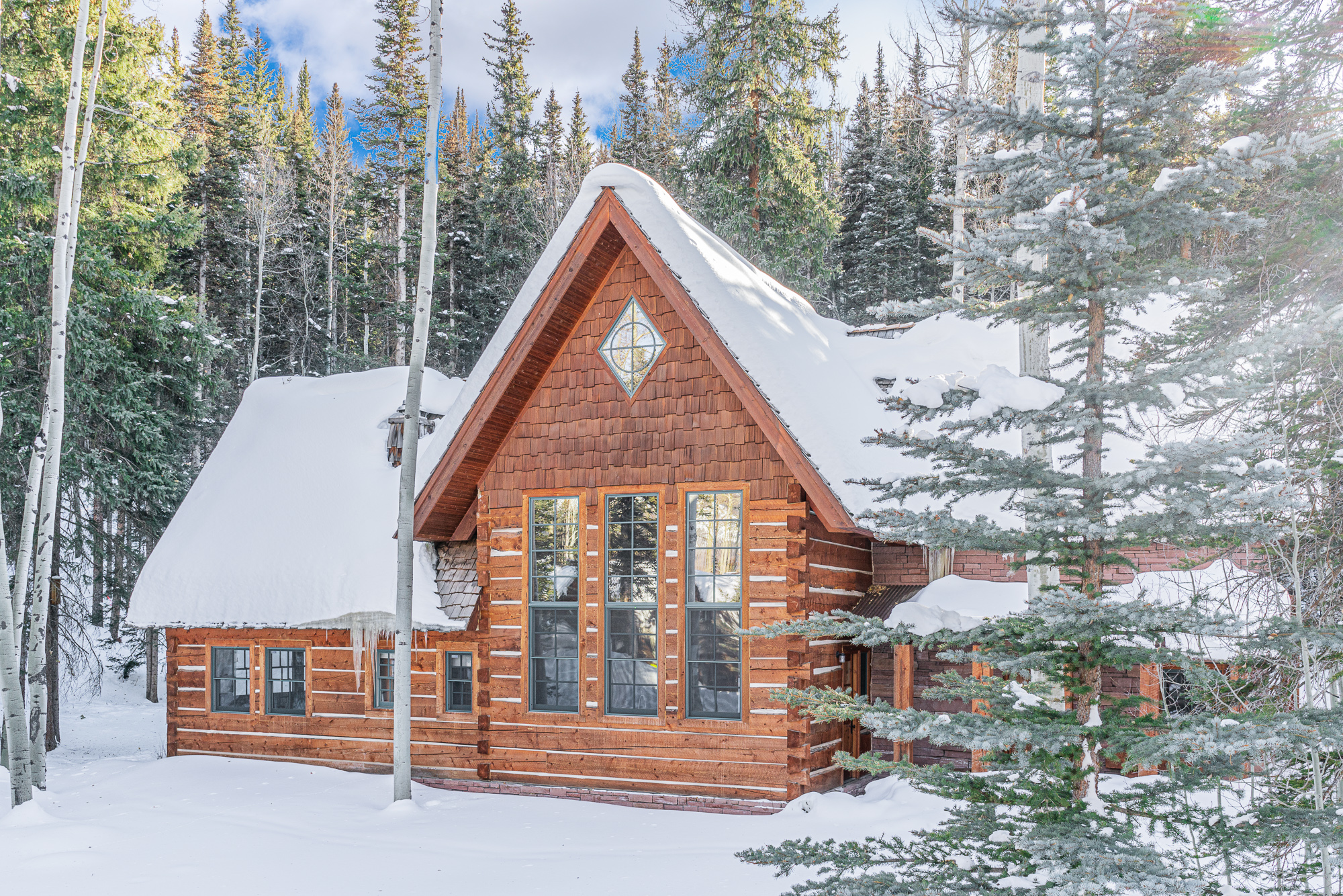 Wooden cabin covered in snow and surrounded by snow covered trees.