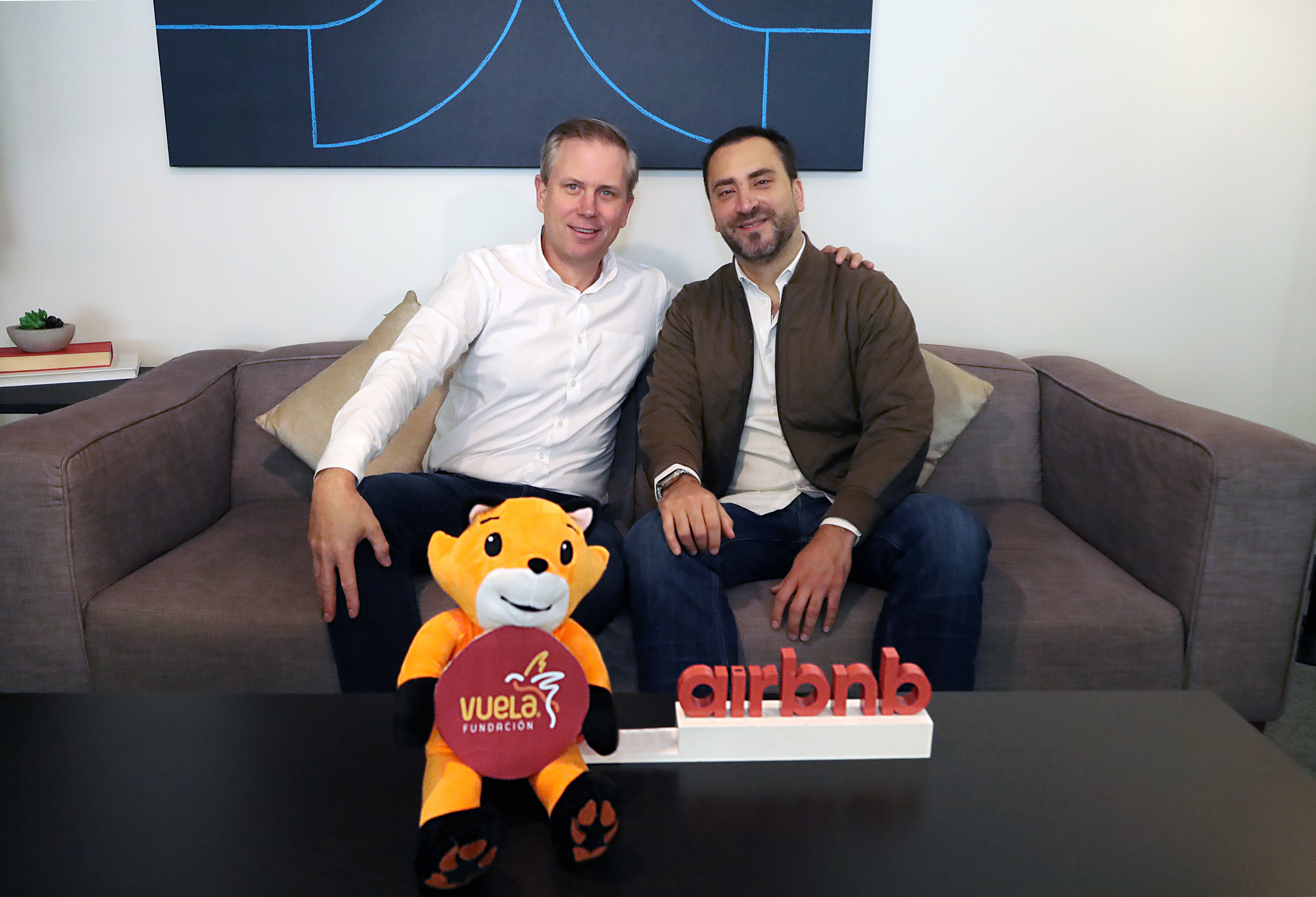 Dos hombres sentados en un sofá, en la mesa de centro el logo de Airbnb y de Vuela