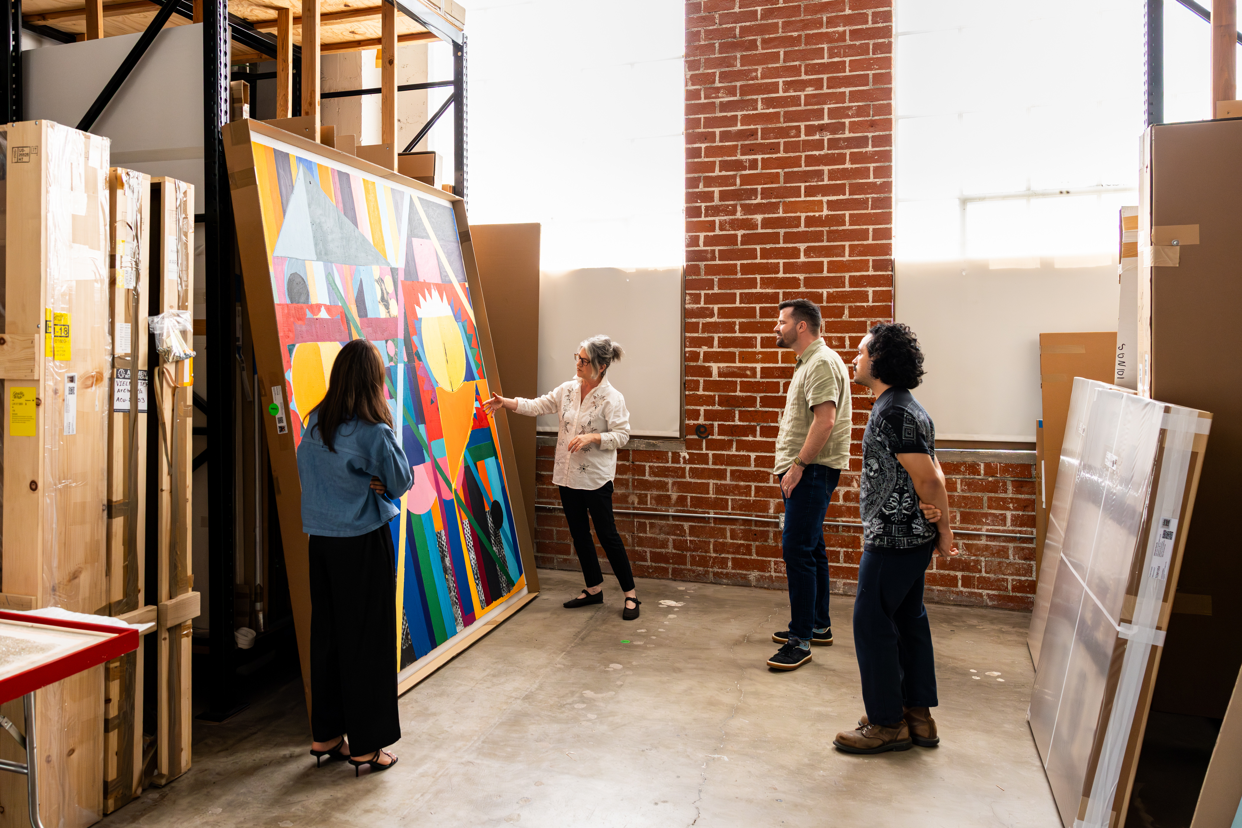 Four people in an art storage room examining a large colorful abstract painting leaning against a wall with brick and white backgrounds.