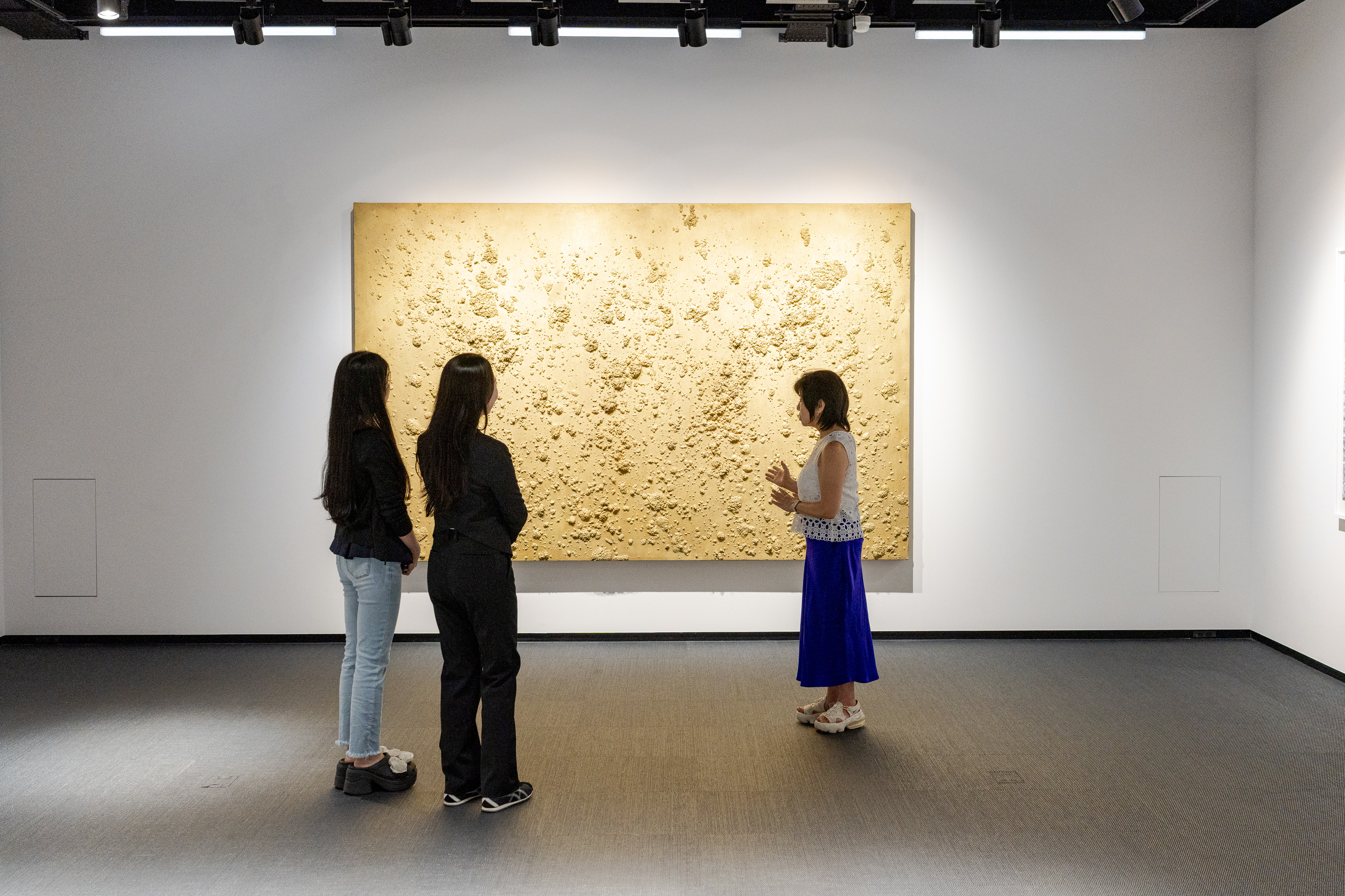 Three women standing in front of a large textured beige-colored artwork in a modern gallery space, discussing the art.
