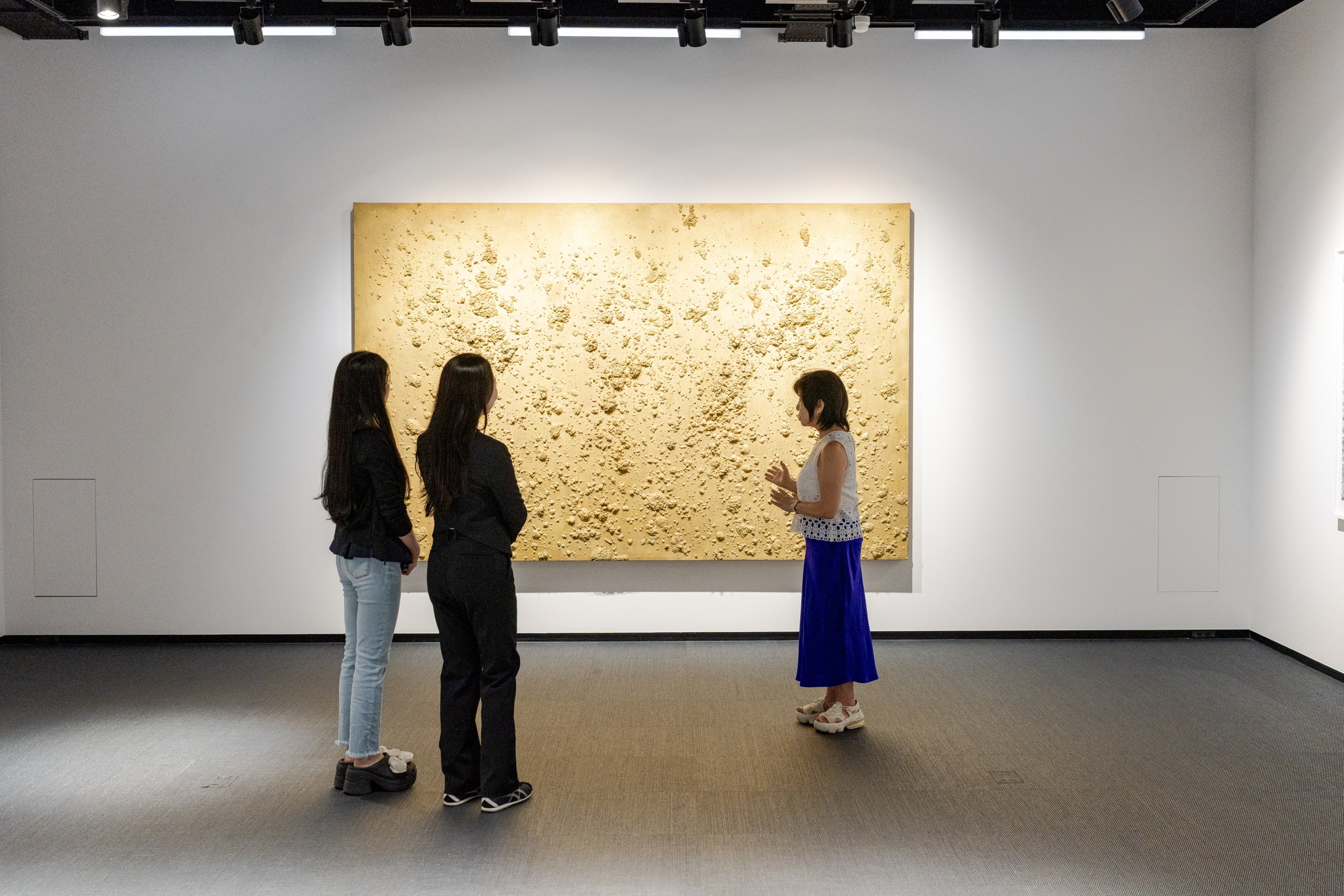 Three women standing in front of a large textured beige-colored artwork in a modern gallery space, discussing the art.