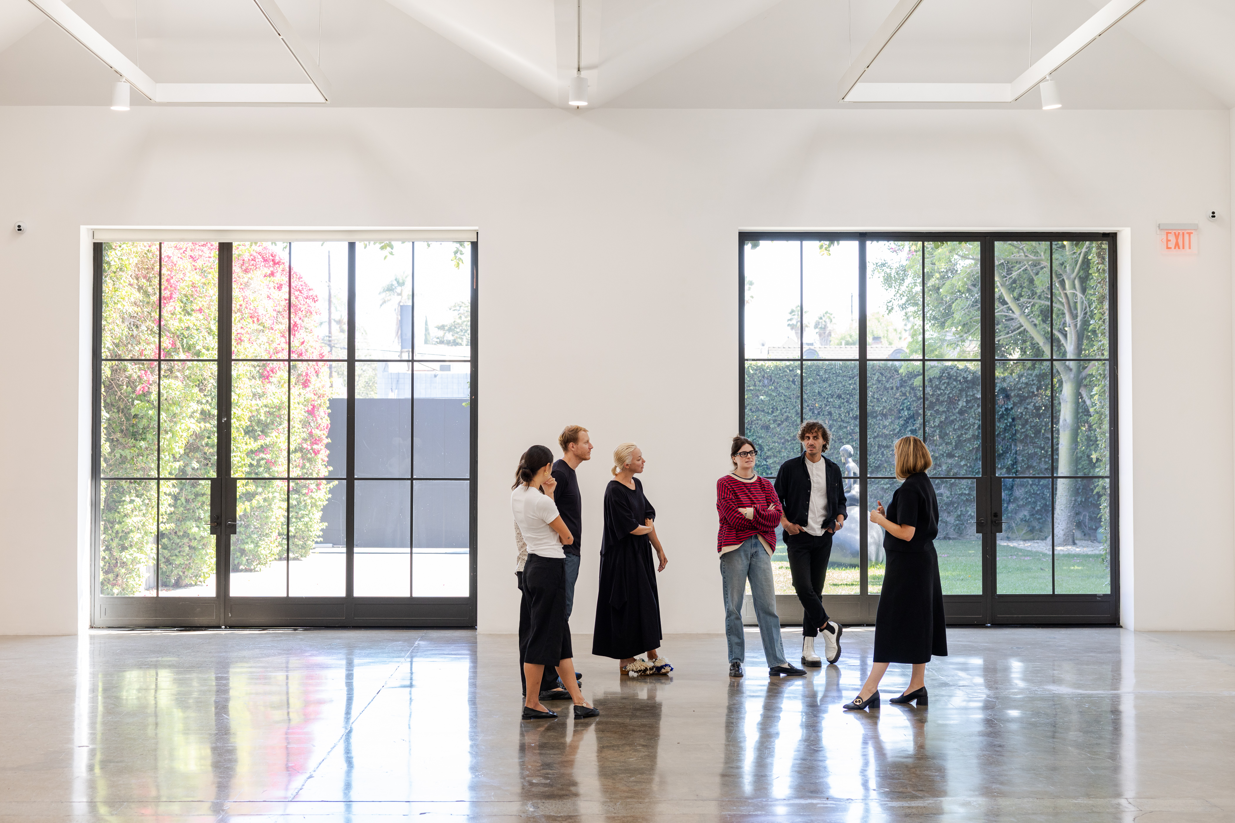 A group of six people standing and talking in a bright, modern space with large floor-to-ceiling windows showing greenery and sunlight outside.