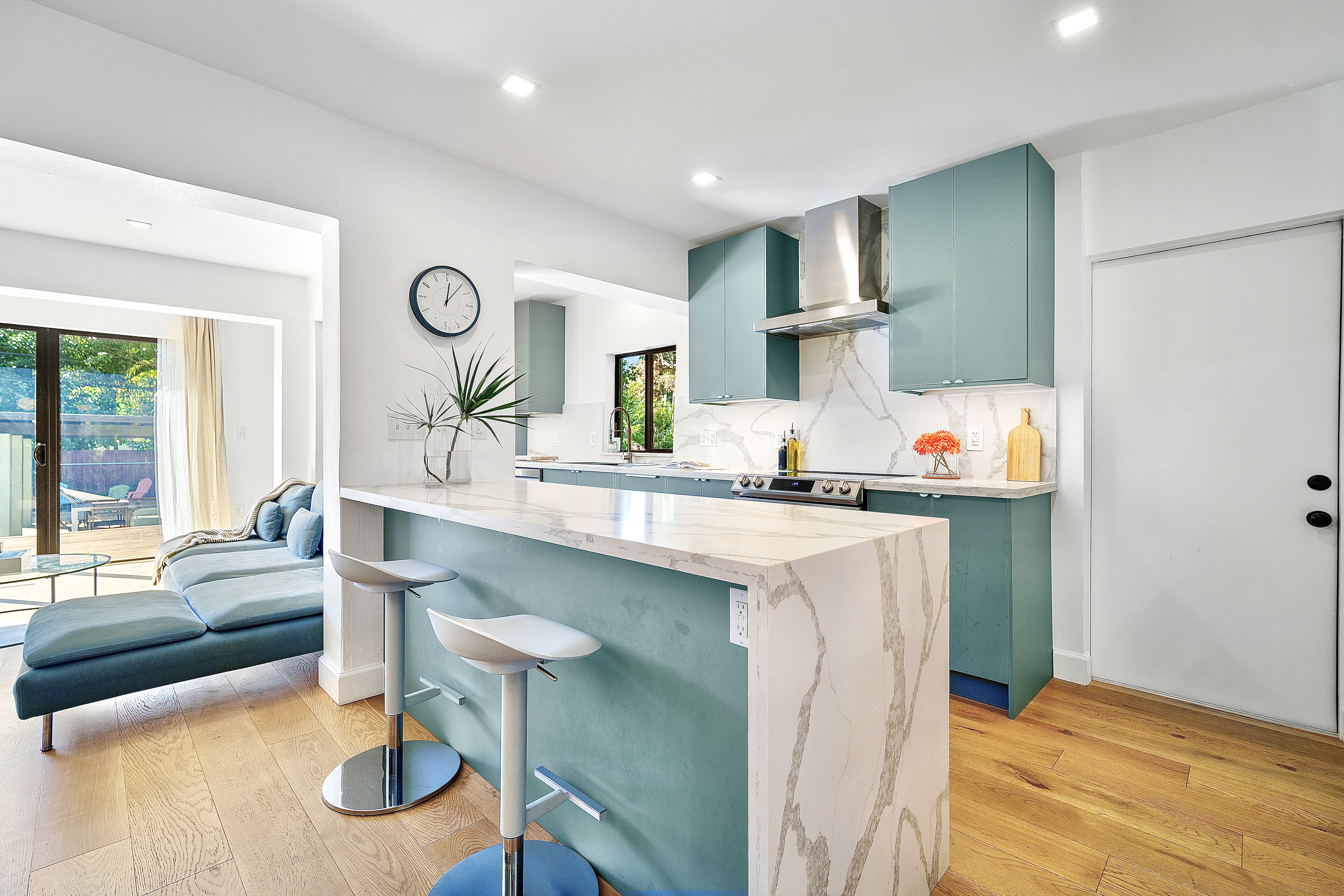 Modern kitchen with teal cabinets, marble countertops, two bar stools, and a view of a living area with a chaise lounge and glass doors leading to an outdoor patio.