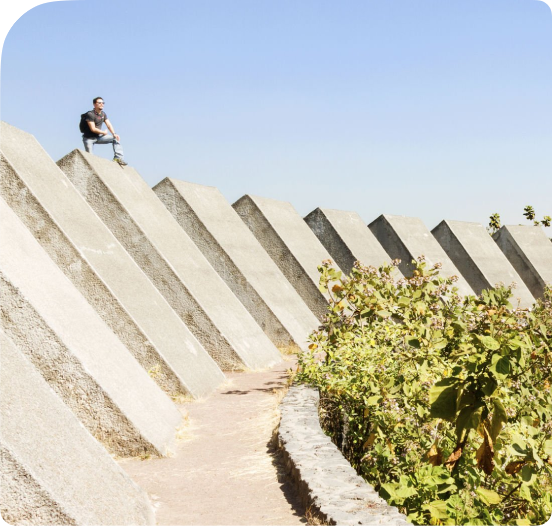 A person sitting atop a series of large, angular concrete blocks with a clear blue sky in the background and greenery along a curved stone pathway in the foreground.