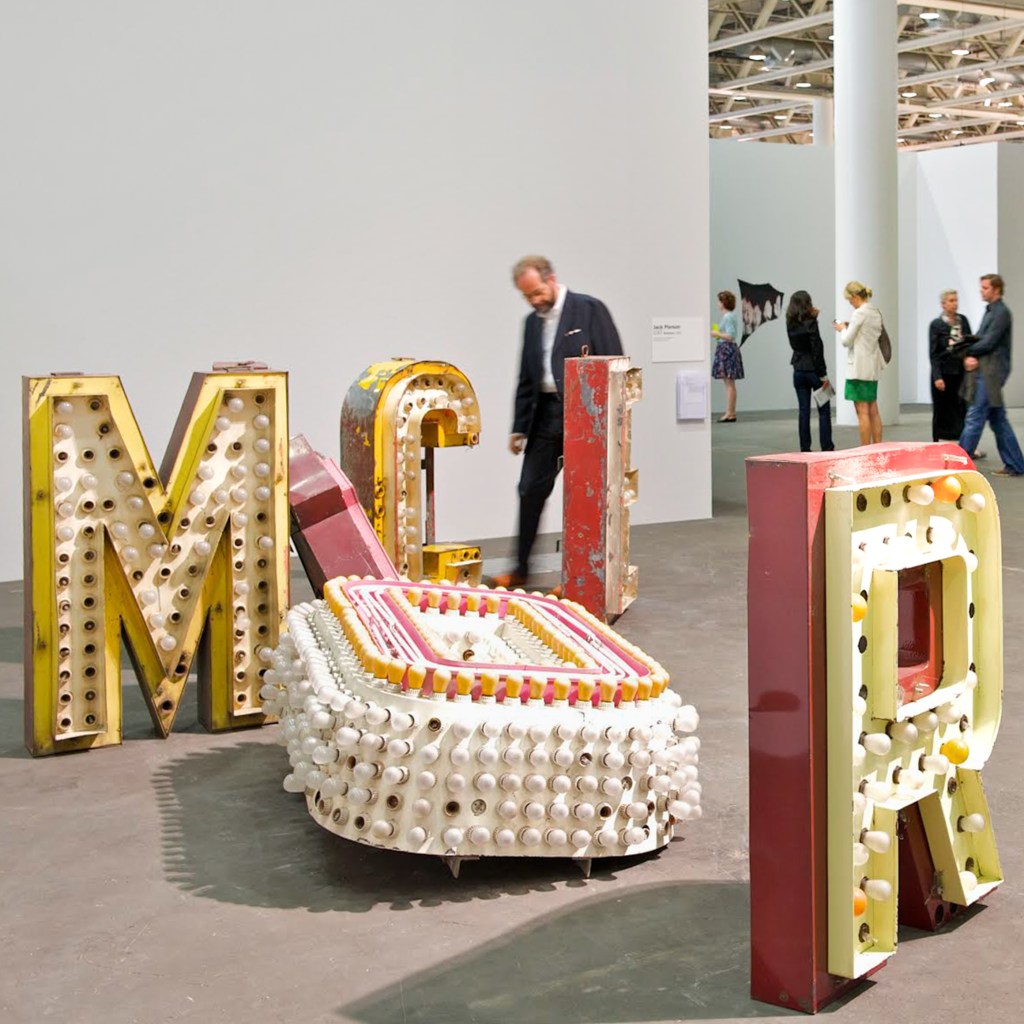 Visitors walk through a spacious art exhibit with bright white walls; in the foreground are colorful sculptures of the letters 'M', 'I', and 'R' made of illuminated bulbs.