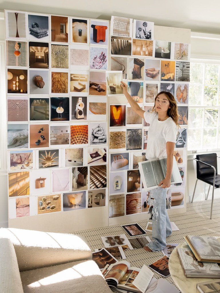 A brightly lit room showcasing a wall covered with a curated collage of design and decor photos, featuring furniture, textures, and artistic elements. A person wearing a casual outfit stands next to the wall, holding an open book and pointing to one of the photos.