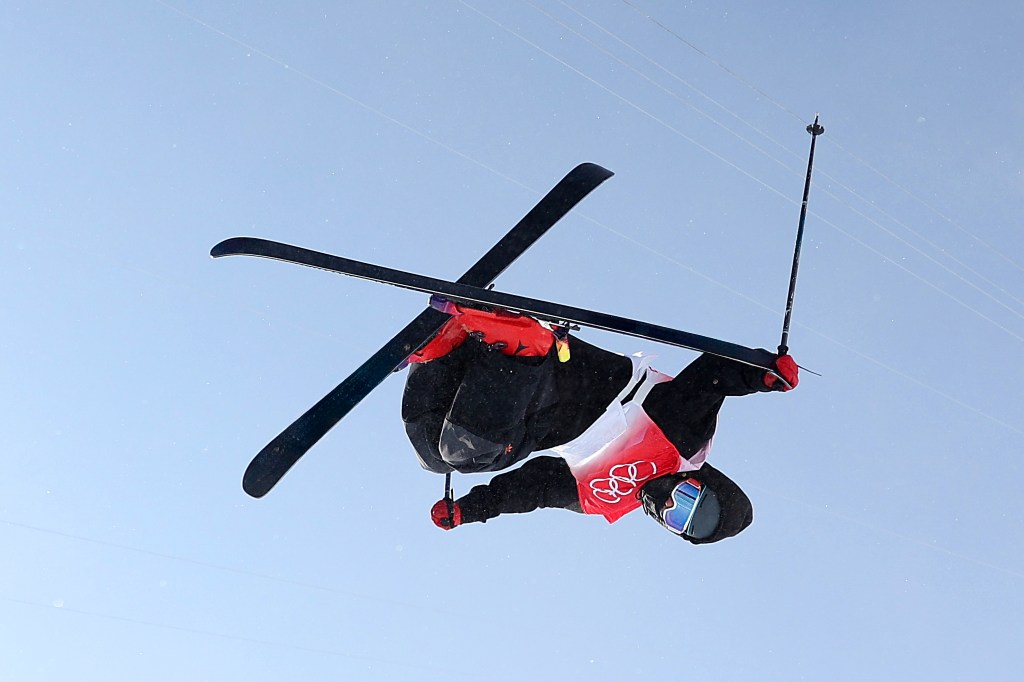 Skier wearing a helmet and reflective goggles mid-air, upside down, executing a freestyle trick with ski poles extended.