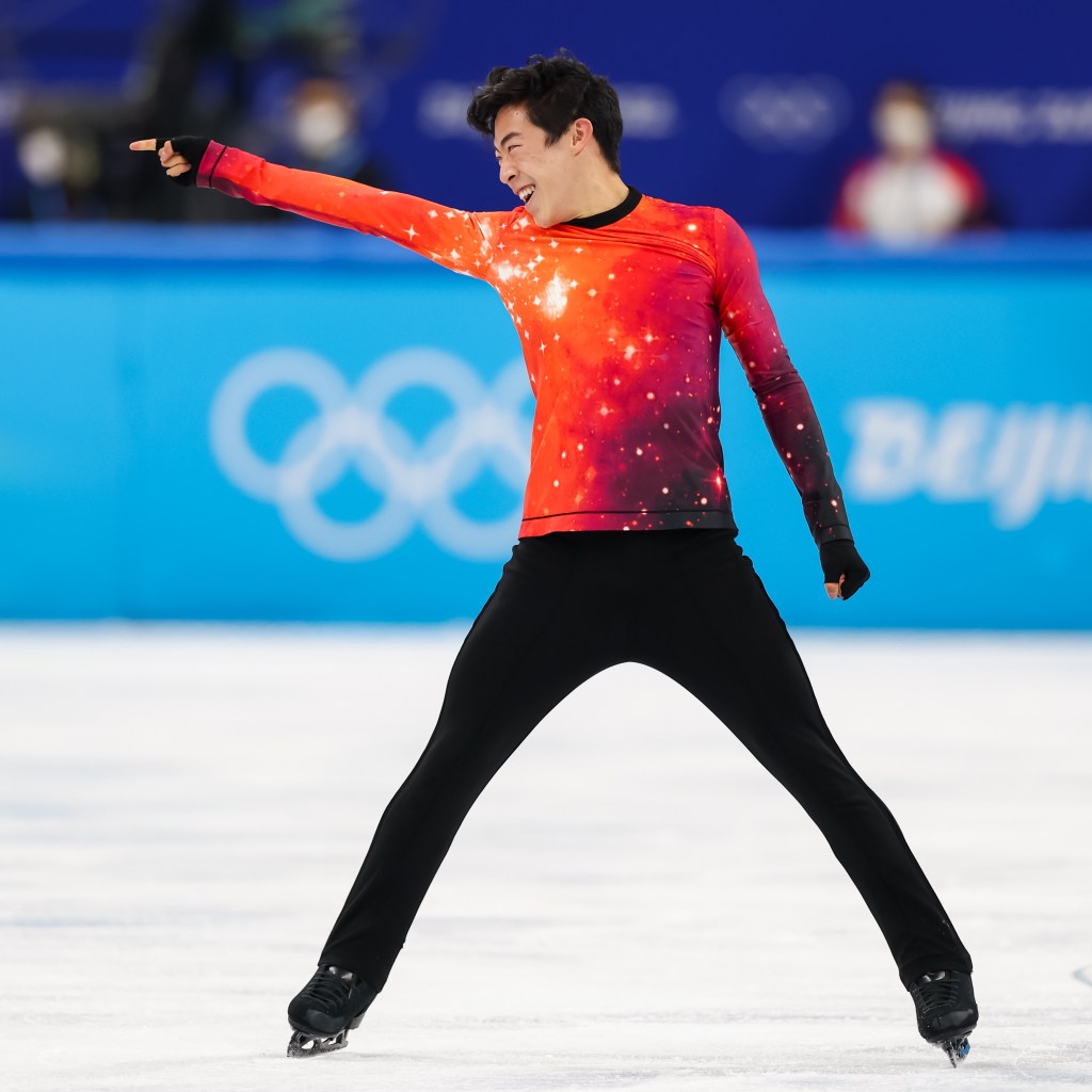 Figure skater performing on ice wearing a vibrant orange and black outfit with galaxy-like patterns, Olympic rings visible in the background.