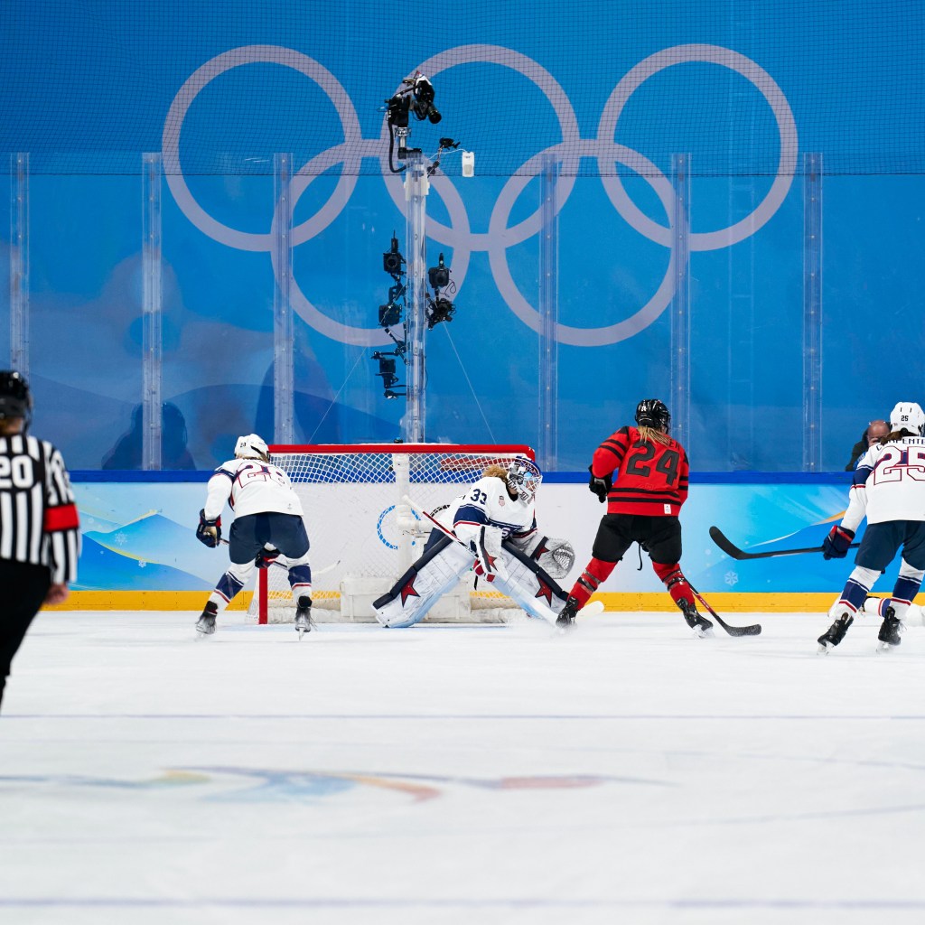 A women's hockey player in a red and black jersey prepares to take a shot at the goal against a goalie in a white and blue jersey during an Olympic match, with the Olympic rings visible in the background.