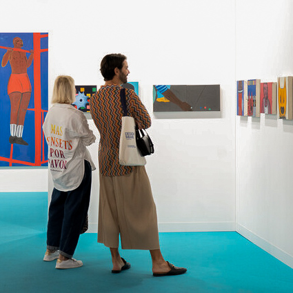 Two people viewing colorful paintings in a modern gallery with bright blue flooring and white walls, including artwork featuring vibrant figures and abstract designs.