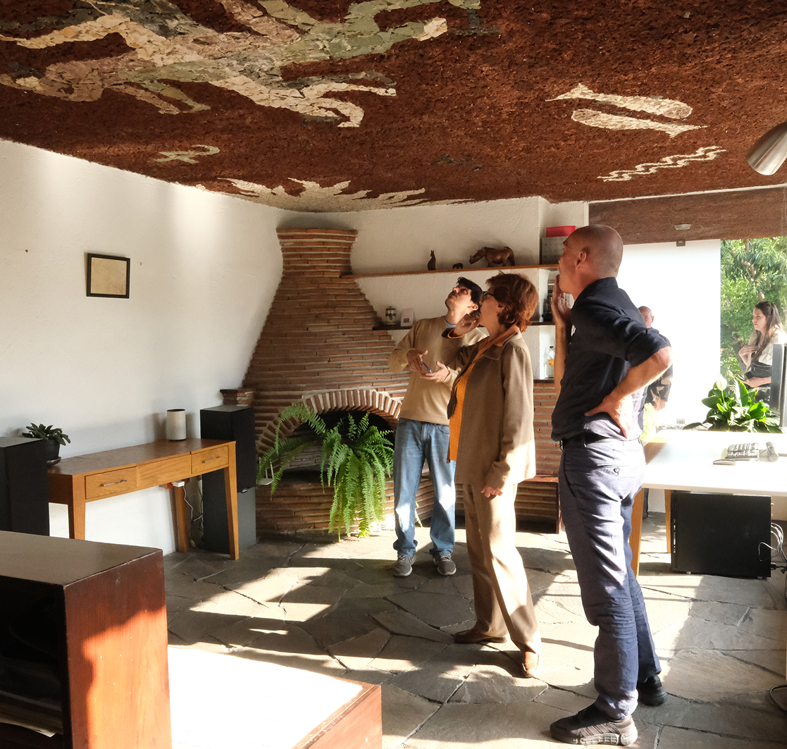 Three people stand in a brightly lit room with a unique textured ceiling featuring abstract designs. One person points upwards toward the ceiling while the others observe. The room has a fireplace, plant decorations, and modern wooden furniture.
