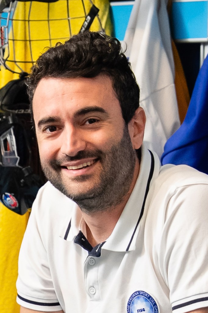 A person wearing a white polo shirt with a blue embroidered logo, seated in front of sports gear including a yellow jersey and hockey equipment.