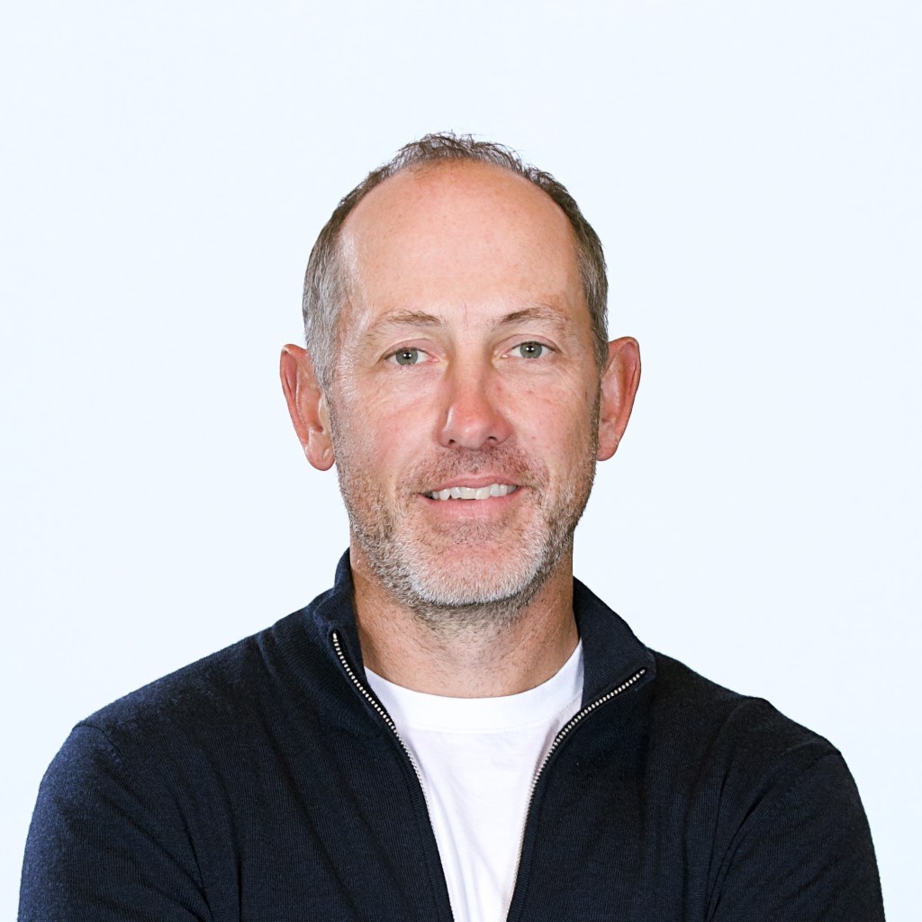 Close-up of an individual dressed in a navy sweater and white undershirt, shown from the shoulders up with arms crossed.